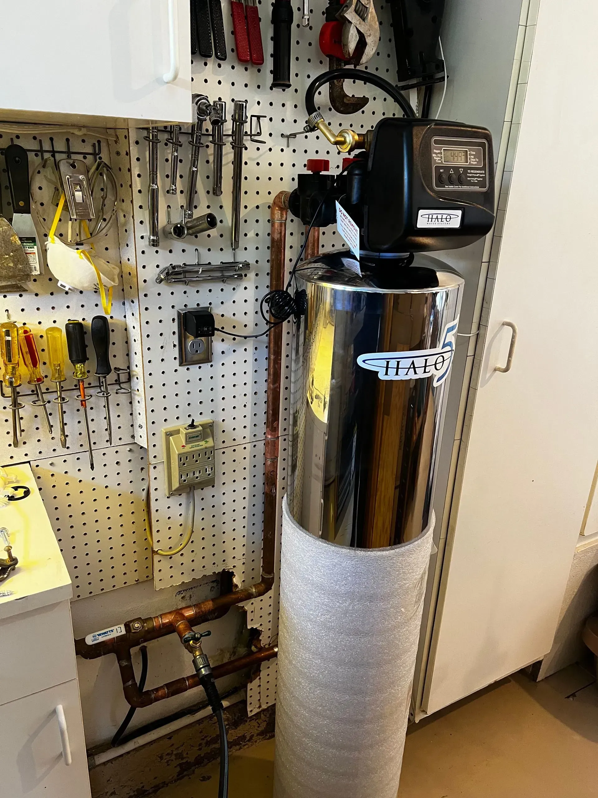 Water softener in a garage, shiny tank wrapped in bubble wrap, copper pipes, tools on a pegboard.