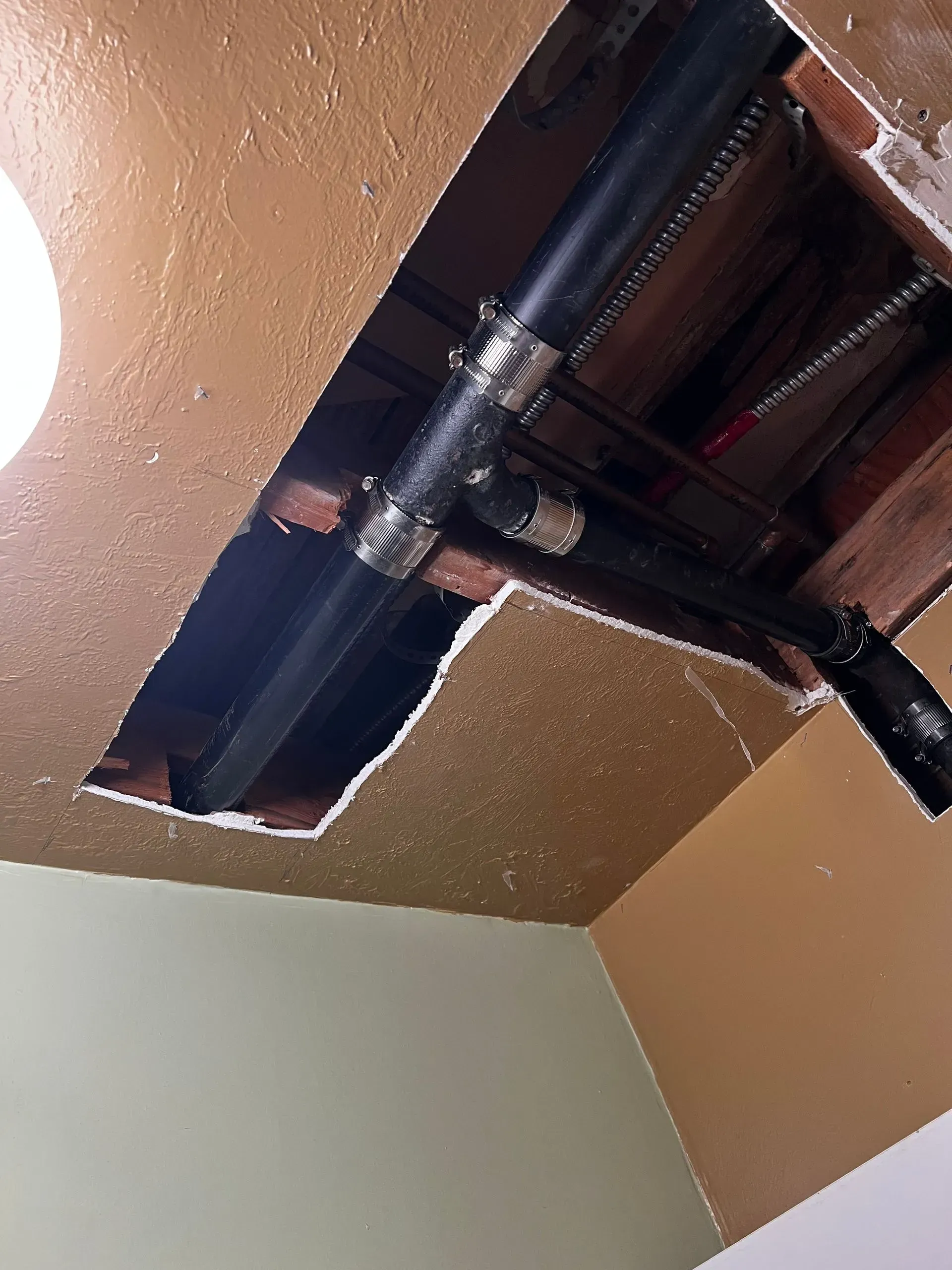 Black plumbing pipes in a damaged ceiling. Metal straps hold pipes together. Brown ceiling and exposed wood framing.