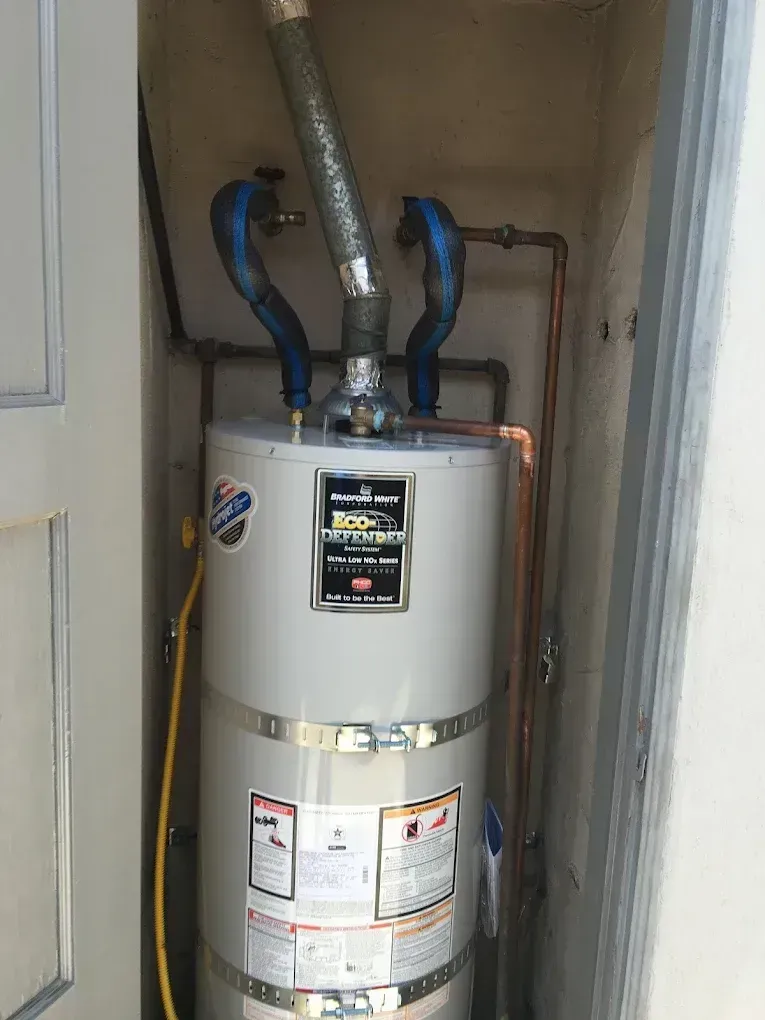 A tall silver water heater with blue pipes and a vent inside a utility closet.
