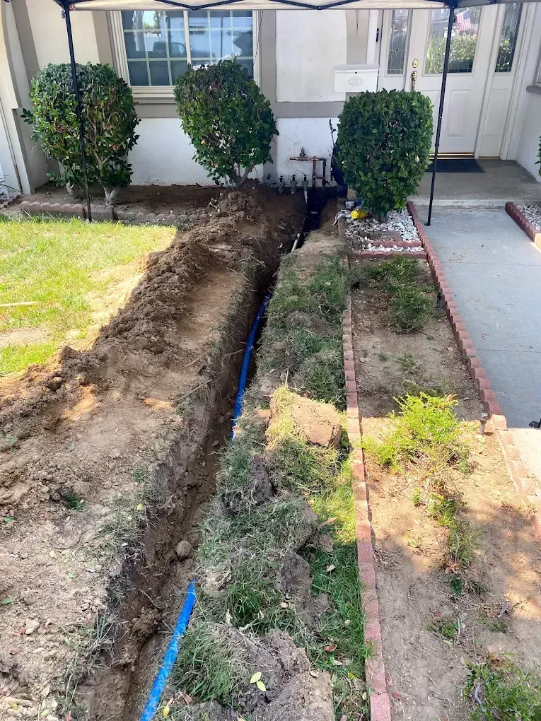A long trench in a yard with blue pipe, bushes, and a house in the background.