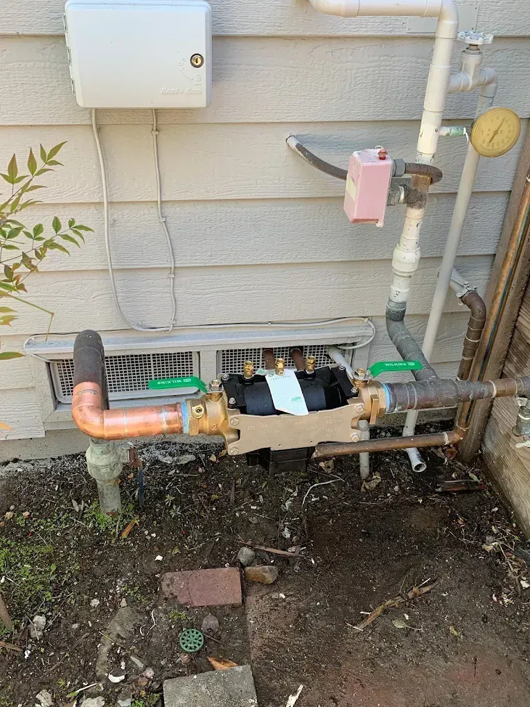 Outdoor water meter assembly with pipes, valves, and a pressure gauge, mounted on a building's exterior.