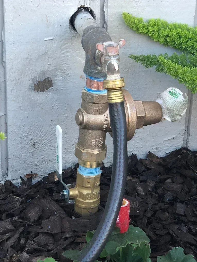 A water valve system with attached hose, mounted on a concrete wall, set in wood chips.