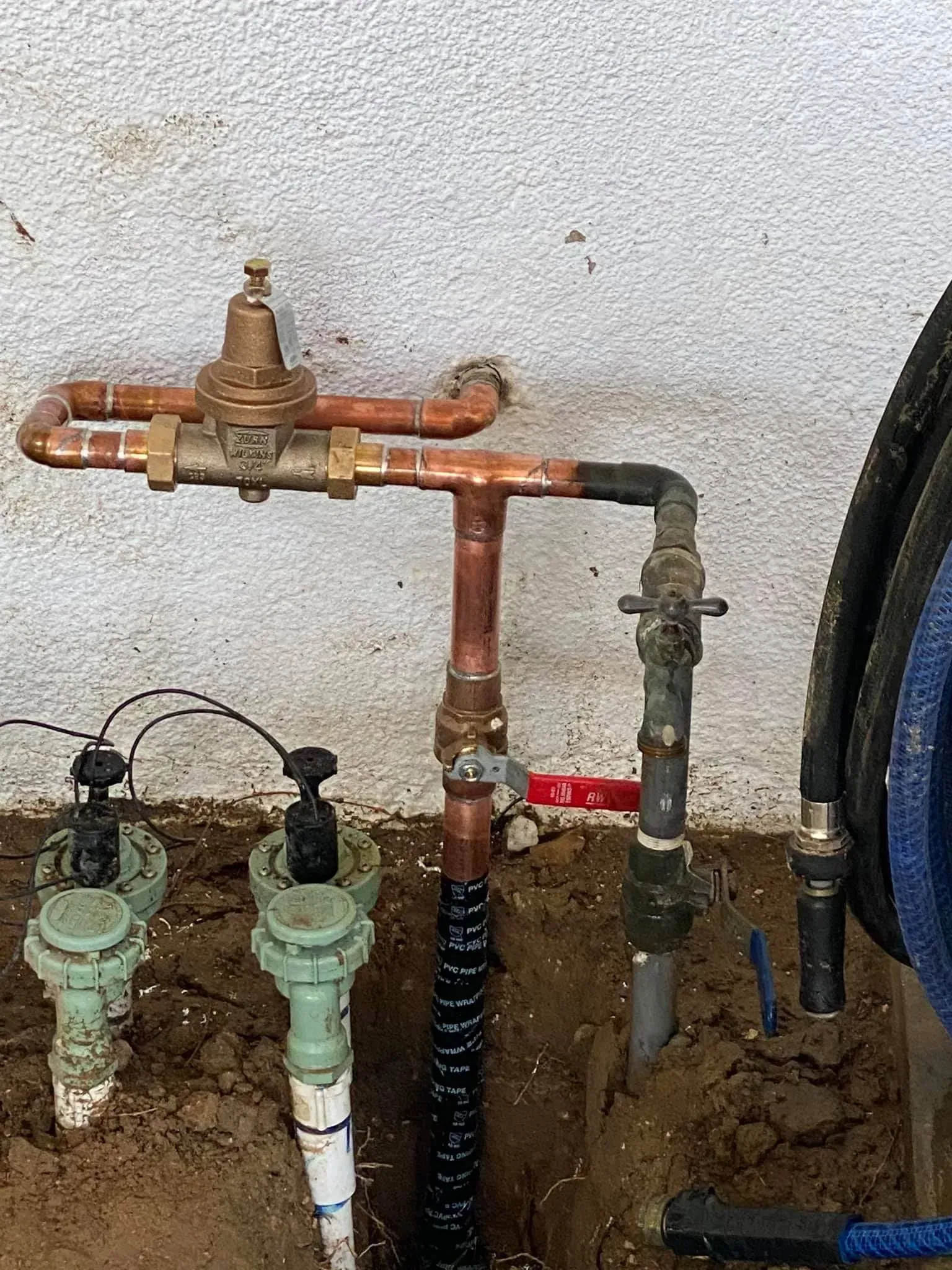 Copper and PVC irrigation system components and sprinkler heads installed near a wall in a dirt setting.