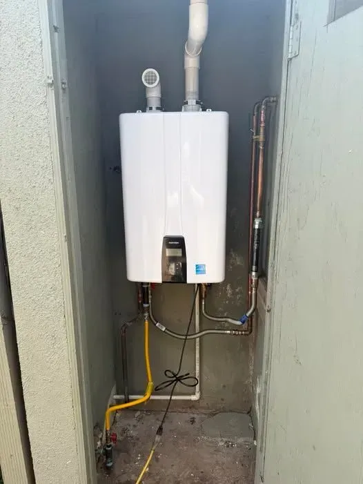 A white tankless water heater installed in an outdoor enclosure, with gas and water lines connected.