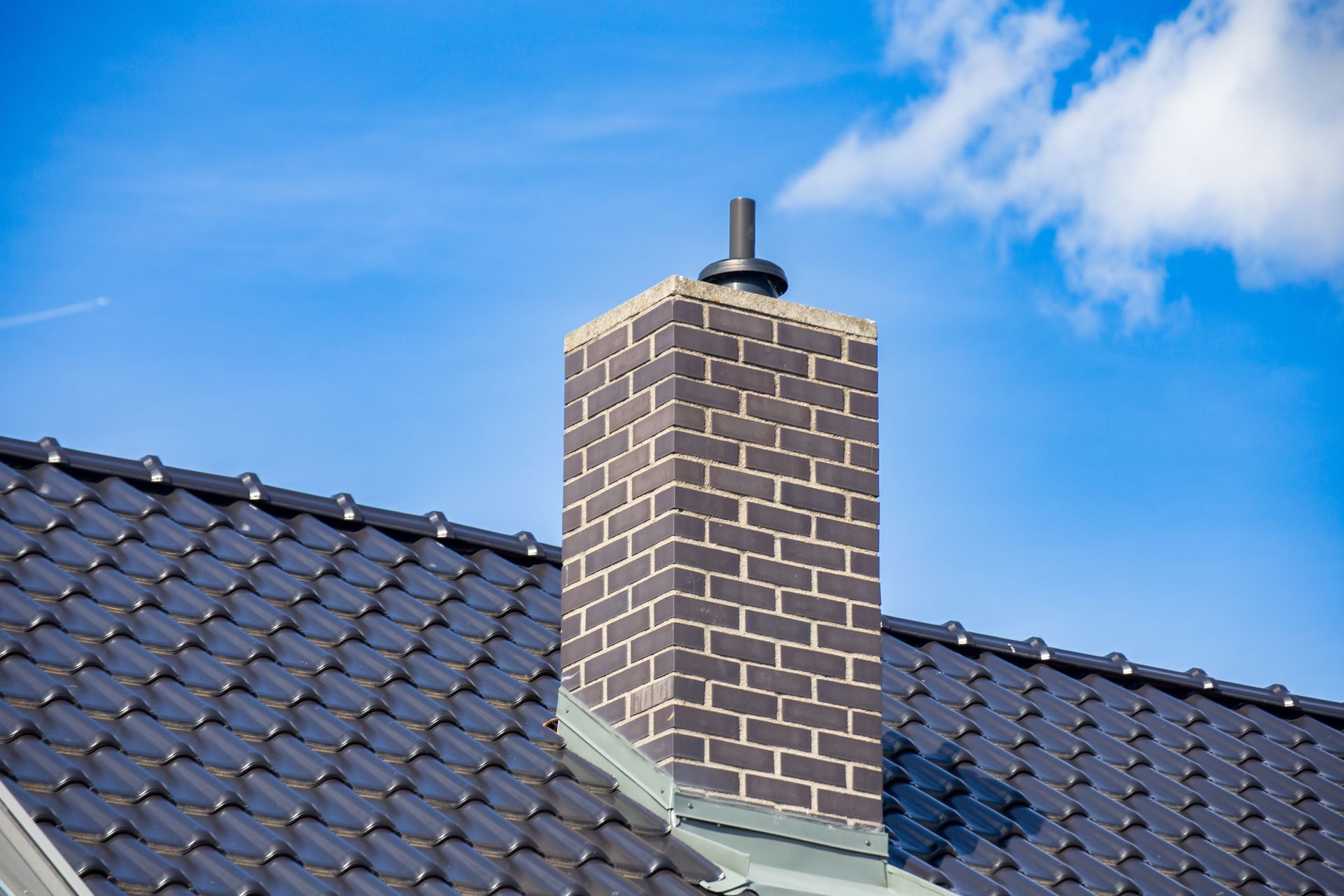 A brick chimney with a black metal cap, situated on a roof covered in glossy, dark, wavy tiles against a blue sky.
