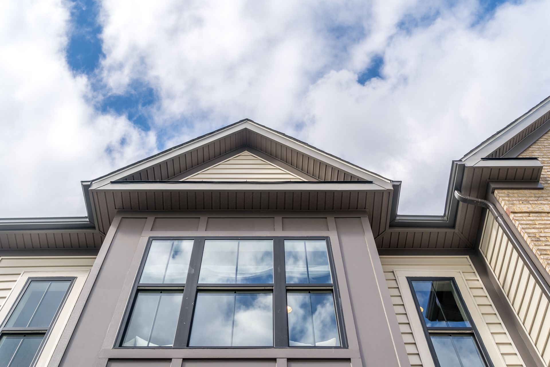 Low-angle view of a beige house exterior with a large window and gable roof against a blue sky with white clouds.