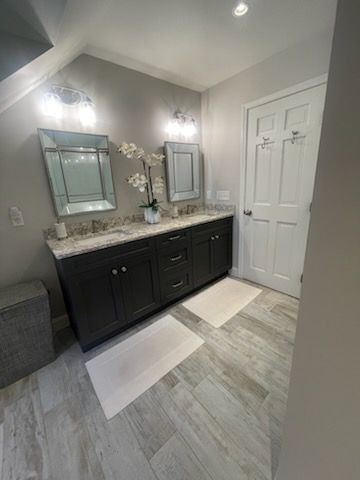 A modern bathroom with a dark double vanity, granite countertops, two wall mirrors, and light wood-look tile flooring.