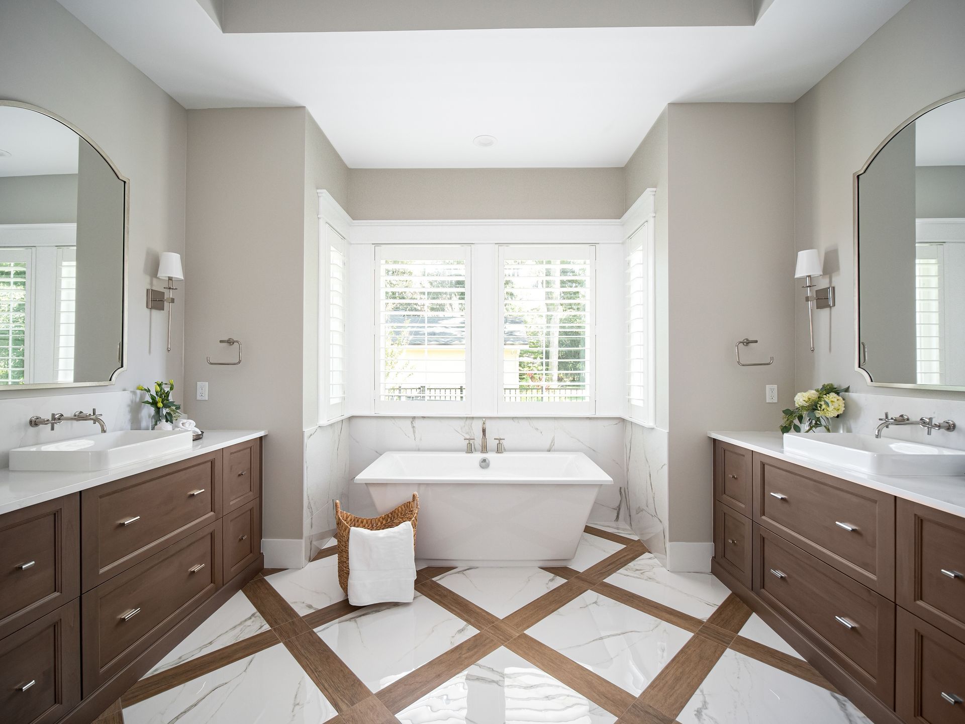 A modern bathroom with two brown vanities, white countertops, arched mirrors, and a freestanding tub below two windows.