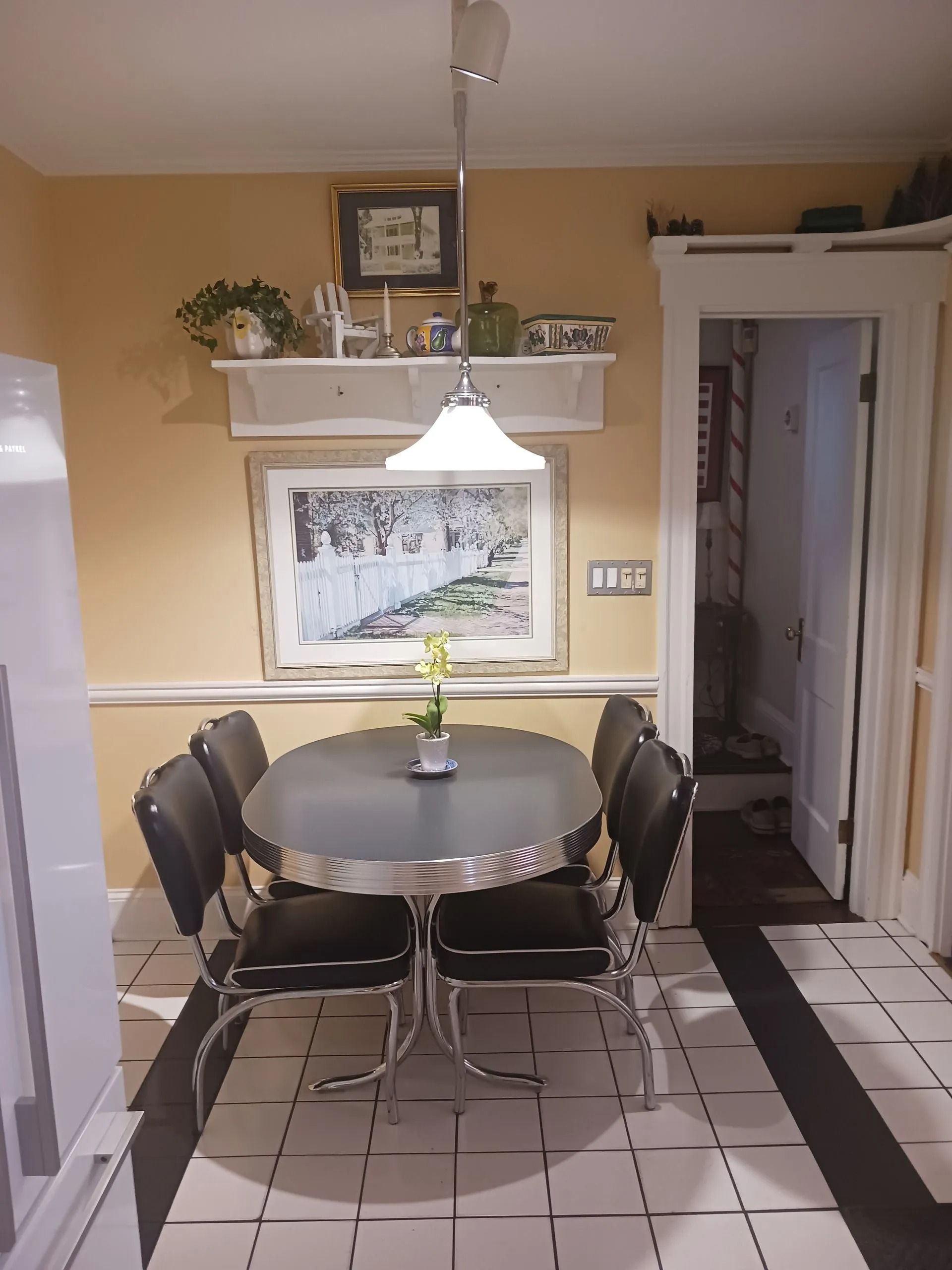 A dining area with a black oval chrome-trimmed table, four black chairs, a wall-mounted shelf, and a framed art print.