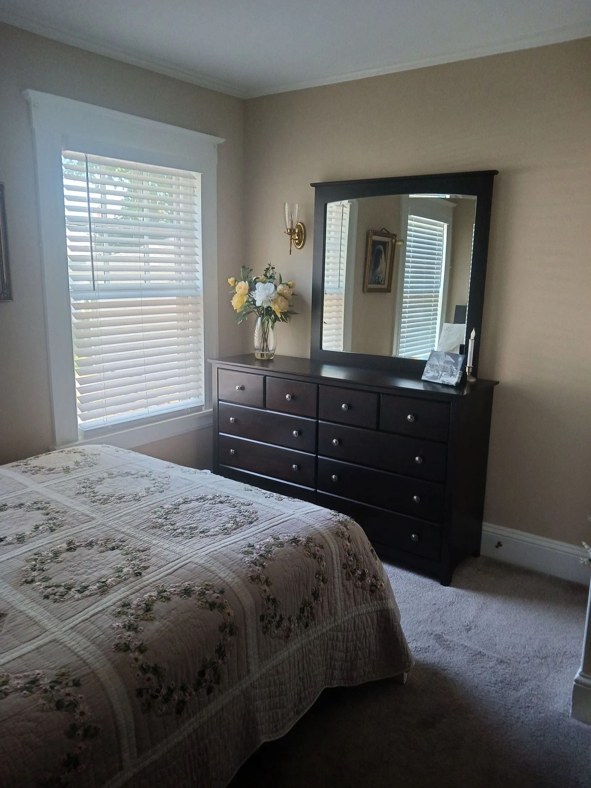 A bedroom with a floral bedspread next to a dark wood dresser with a large mirror and a vase of flowers.