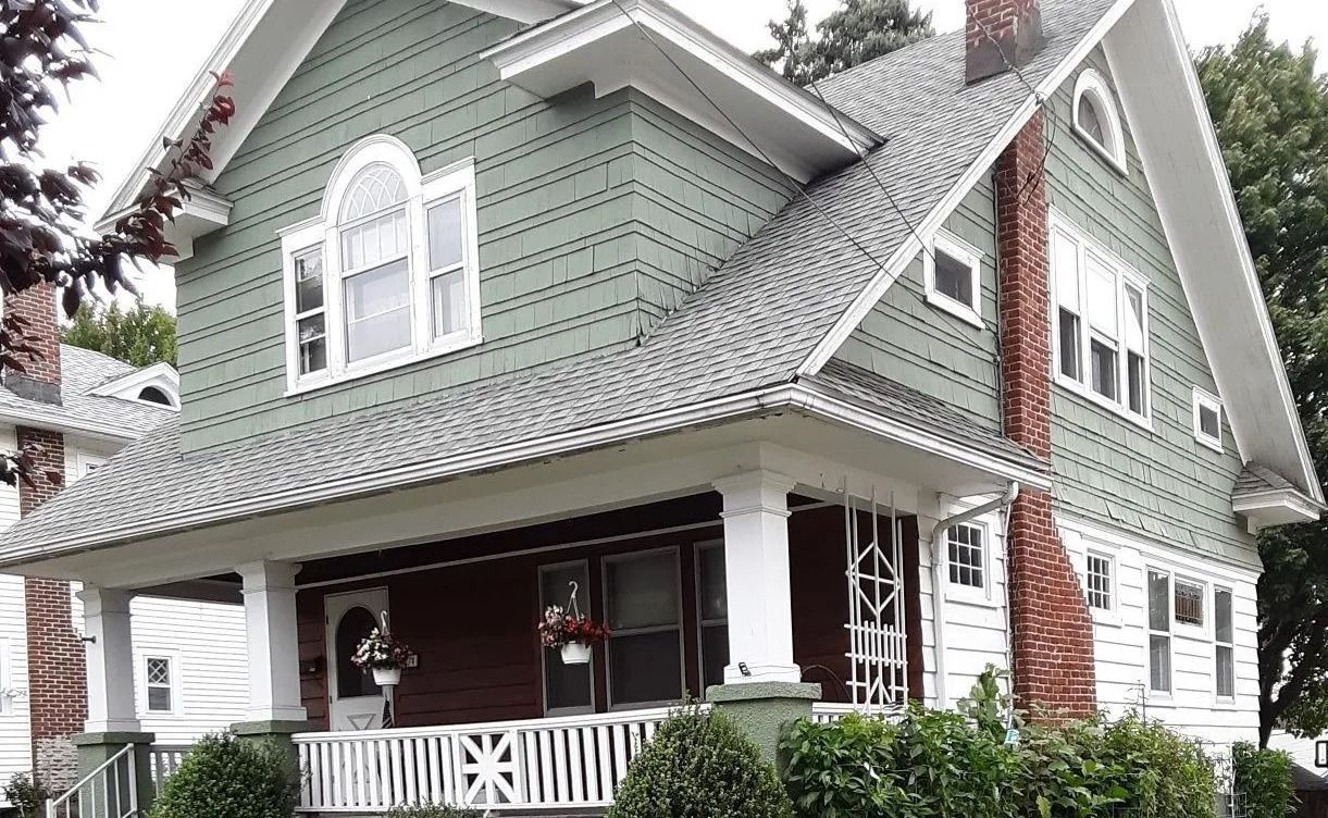 A sage-green two-story Victorian house with white trim, a covered porch, and a brick chimney on a clear day.