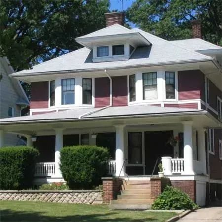 A two-story dark red house with a large white covered front porch, a dormer window, and a stone retaining wall.