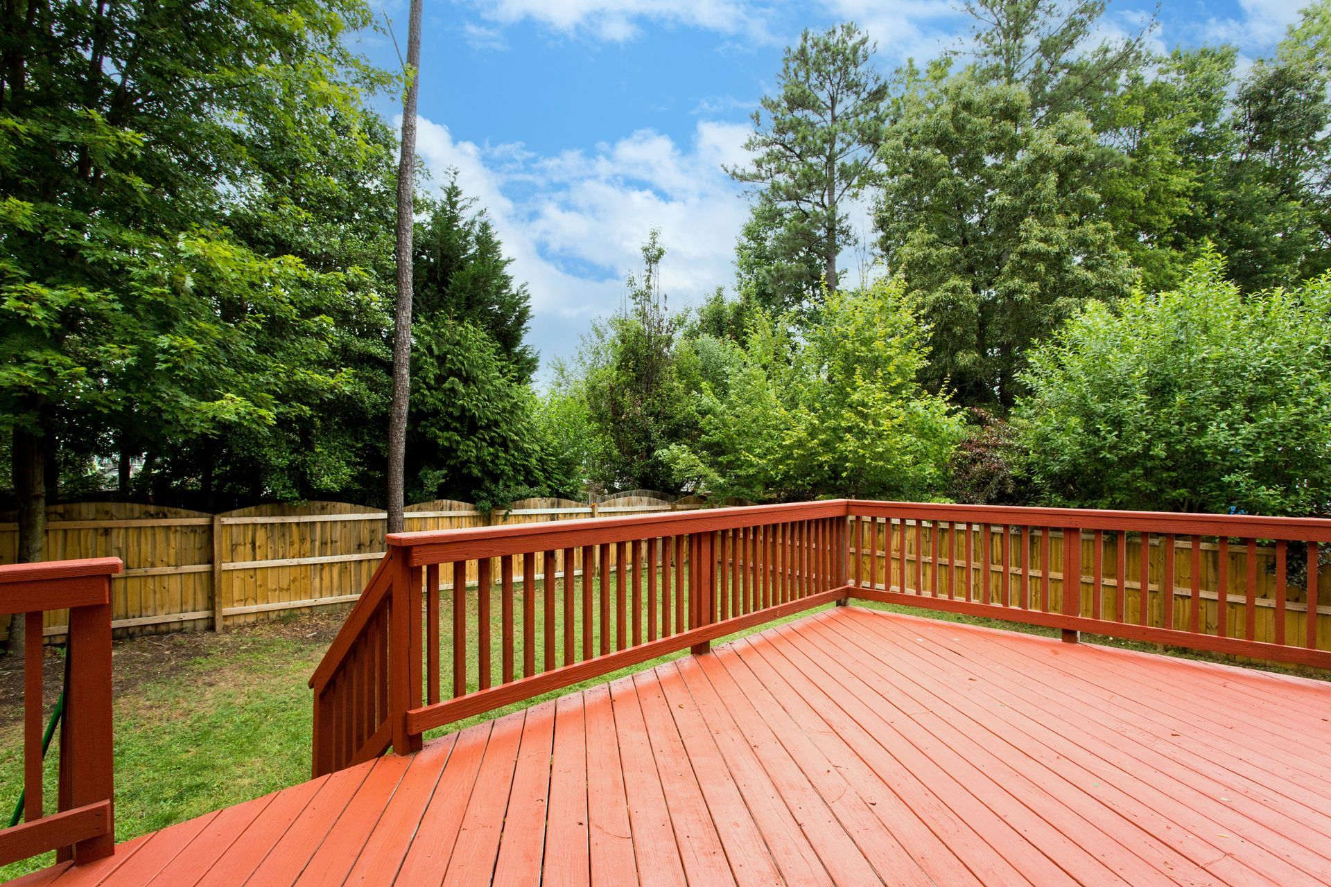 A reddish-brown wooden deck with railings overlooks a backyard with a wooden fence and lush green trees under a blue sky.