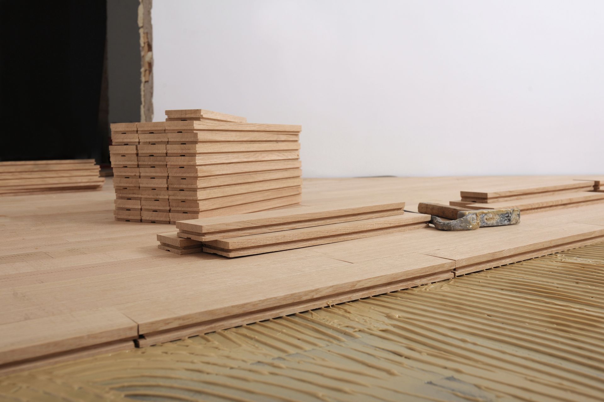 A stack of unfinished wood flooring planks sits on a subfloor prepared with troweled adhesive for installation.
