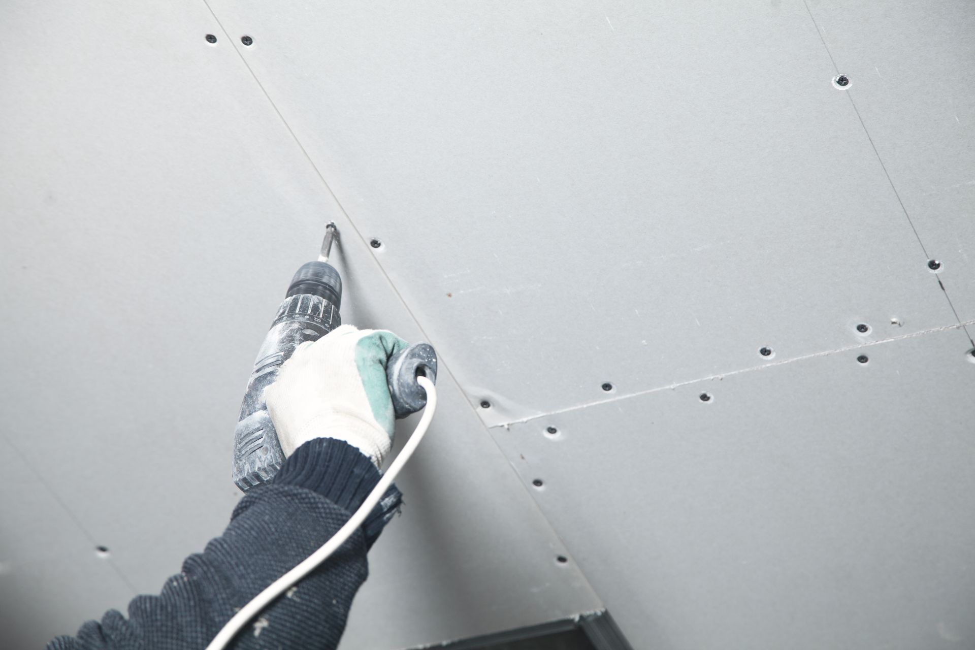 A person uses a power drill to fasten drywall panels to a ceiling.