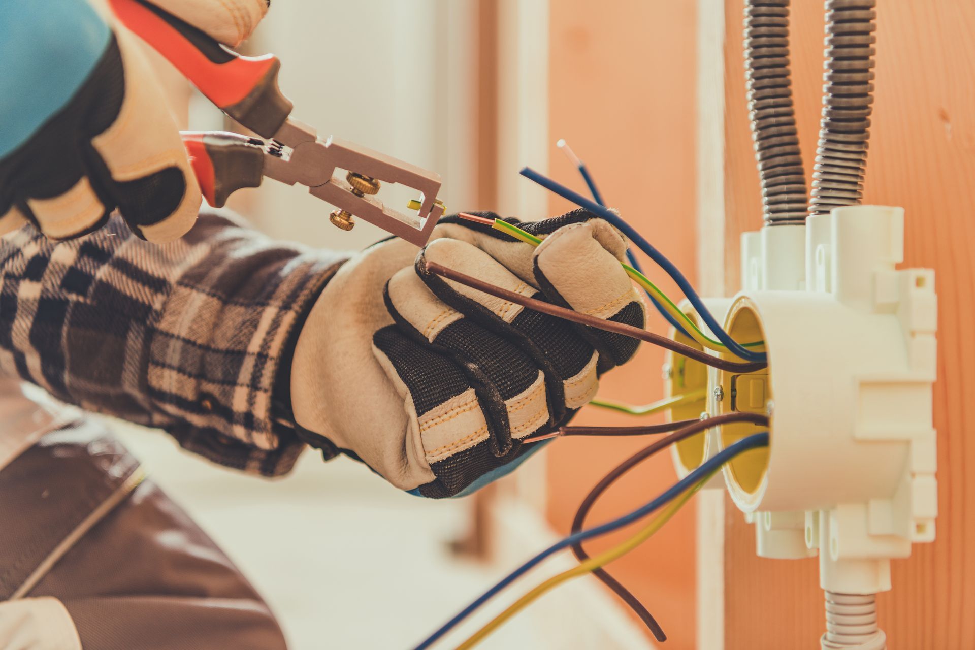 A gloved hand uses wire strippers on electrical wires connected to a white junction box mounted on a wooden wall.