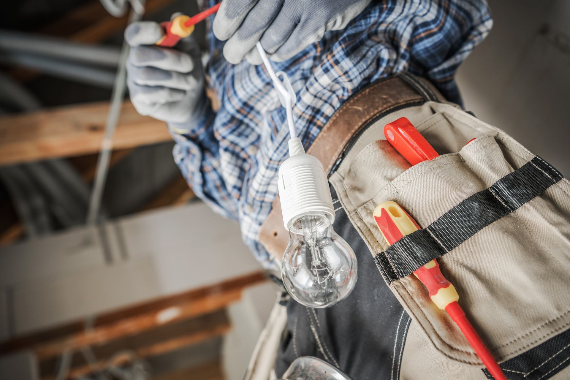 An electrician in a plaid shirt and tool belt holds a light bulb while working on wiring in a construction setting.