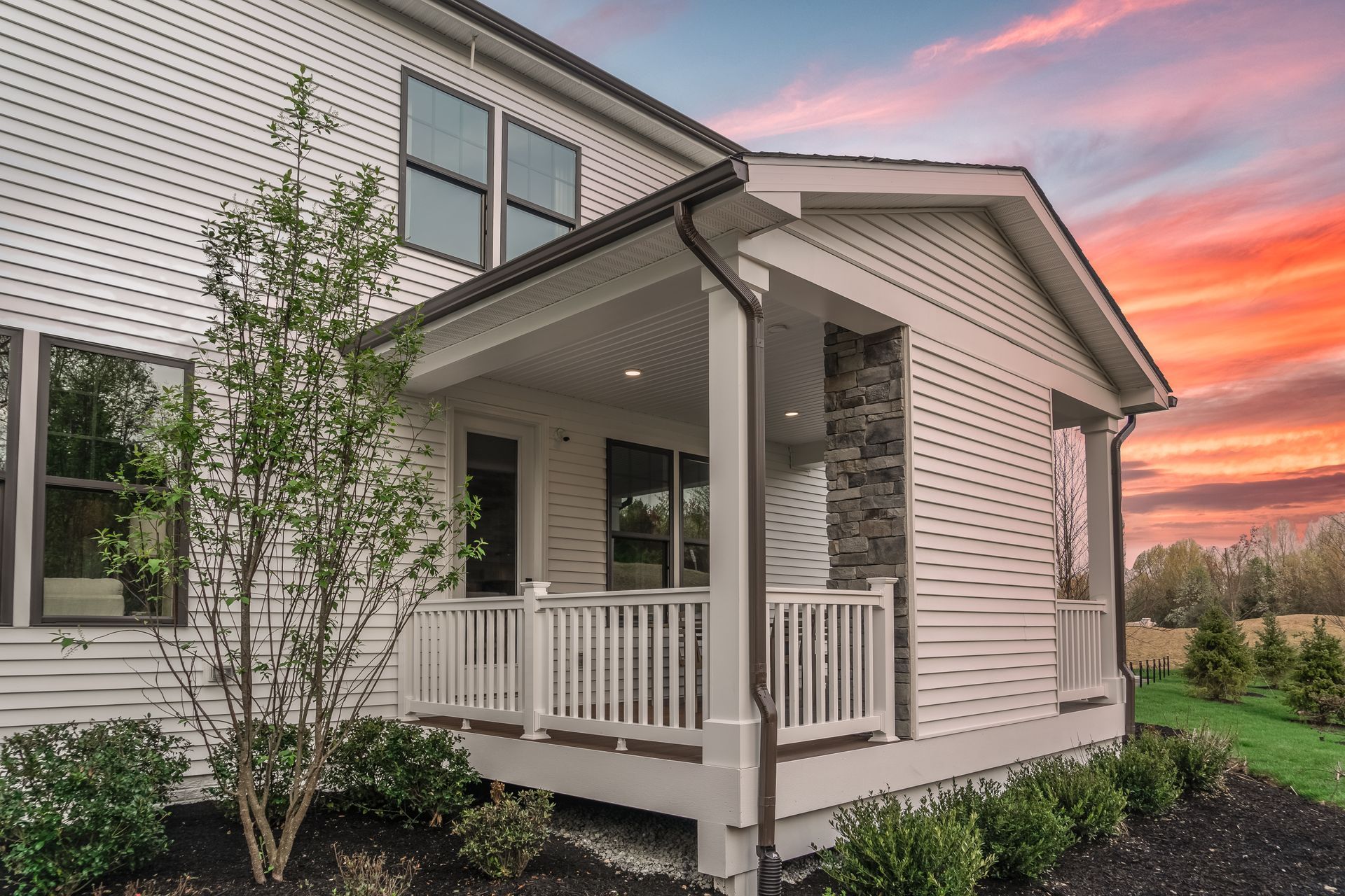 A house exterior featuring a covered porch with white railing, stone accents, and light siding against a sunset sky.