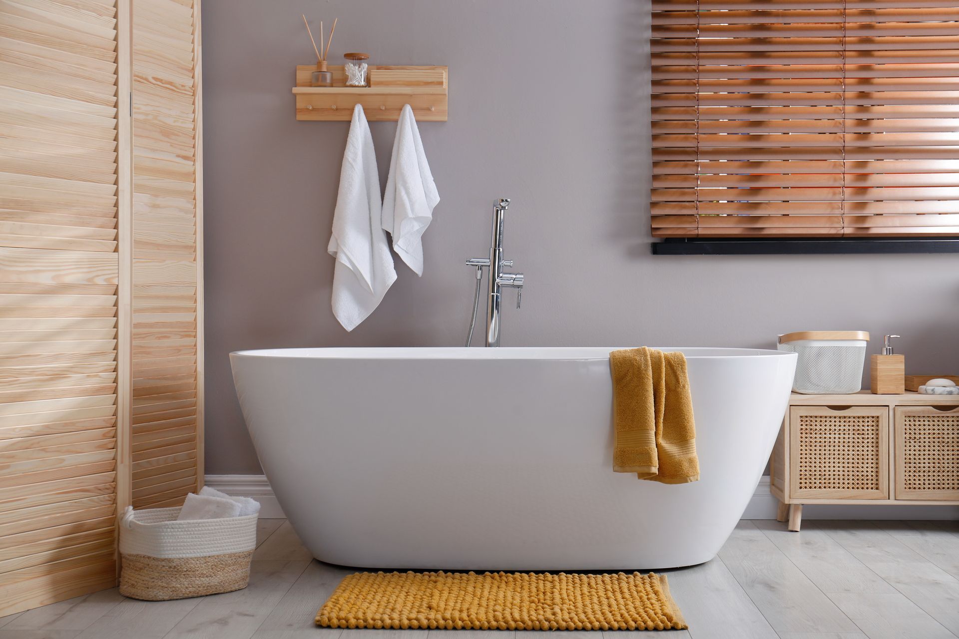 A white freestanding bathtub in a modern bathroom with a wood-slat room divider, yellow bath mat, and hanging towels.
