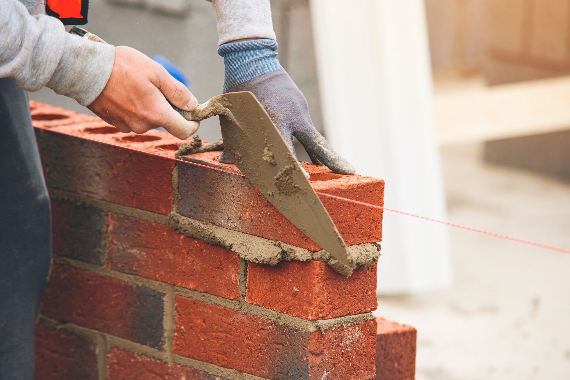 A mason in work gloves uses a metal trowel to spread mortar onto a red brick wall, guided by a horizontal string line.