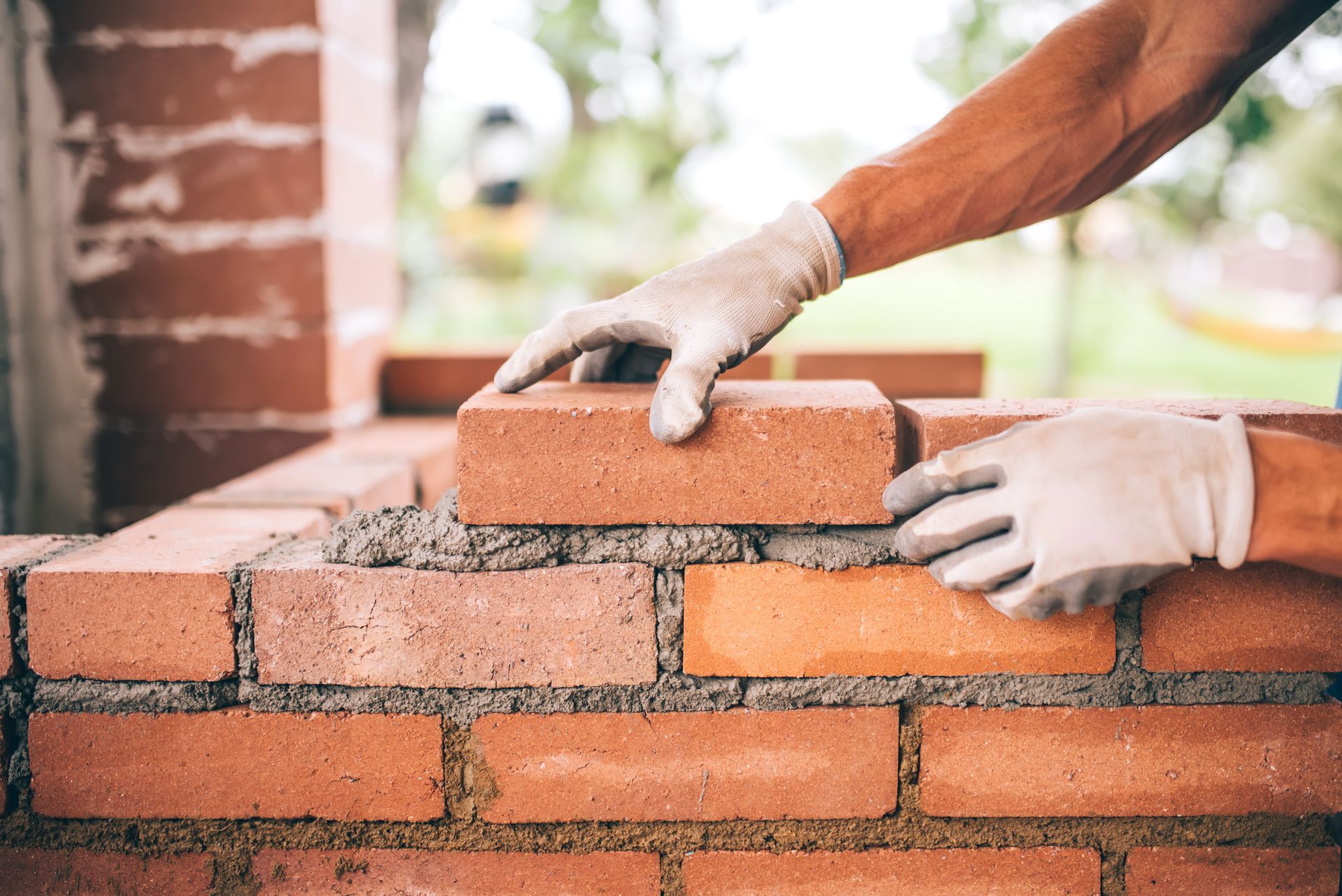 Hands in work gloves placing a red brick onto a fresh layer of mortar during a bricklaying project.