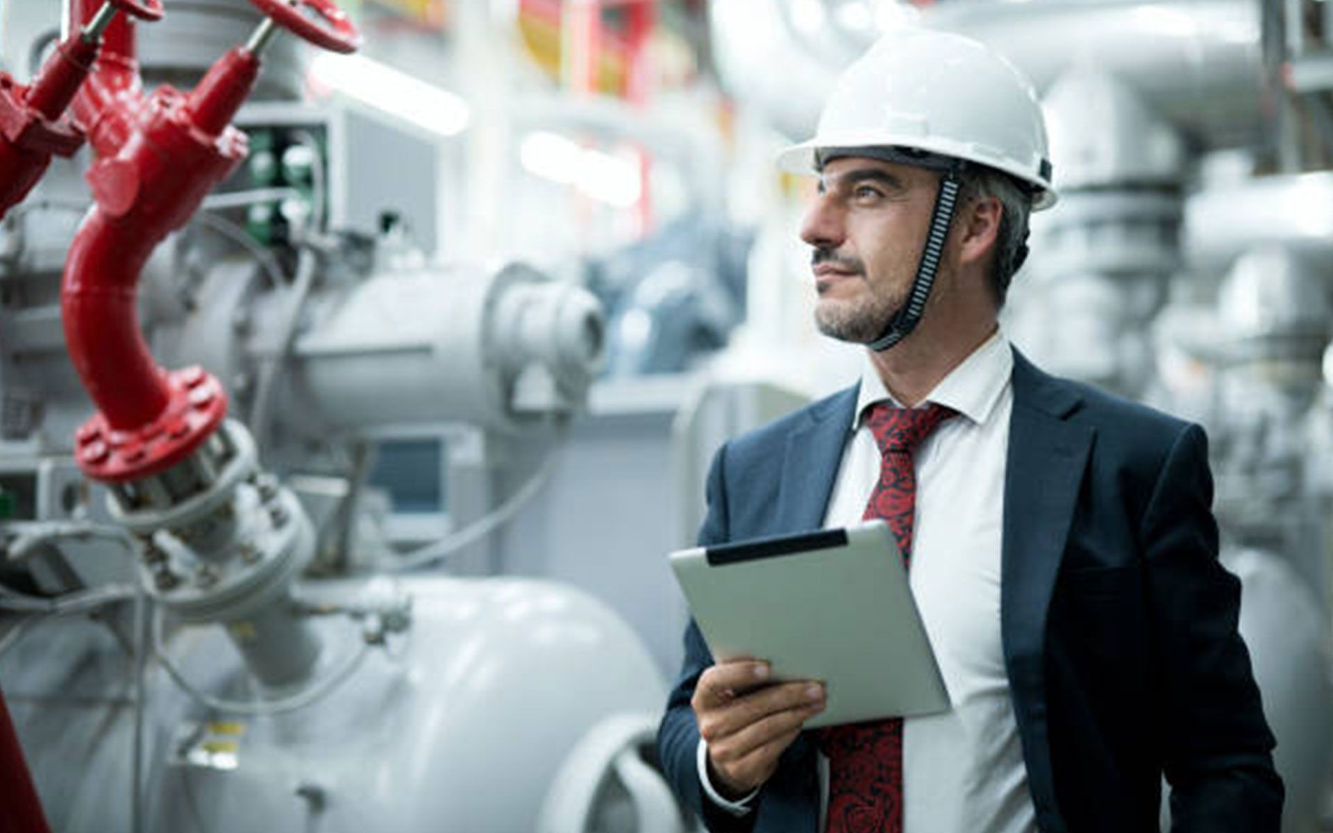 A man in a suit and tie is holding a tablet in a factory.
