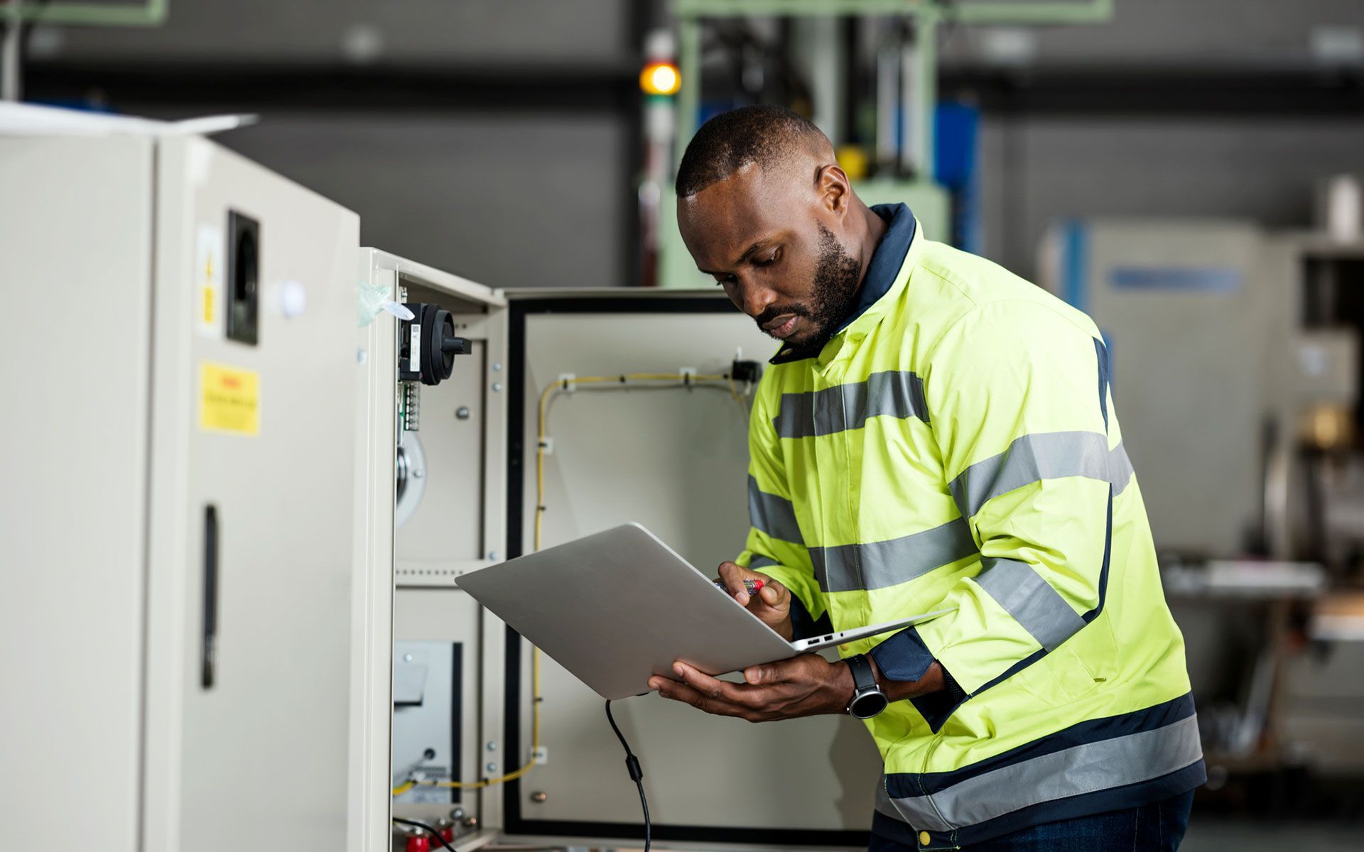A man in a yellow jacket is looking at a clipboard in a factory.