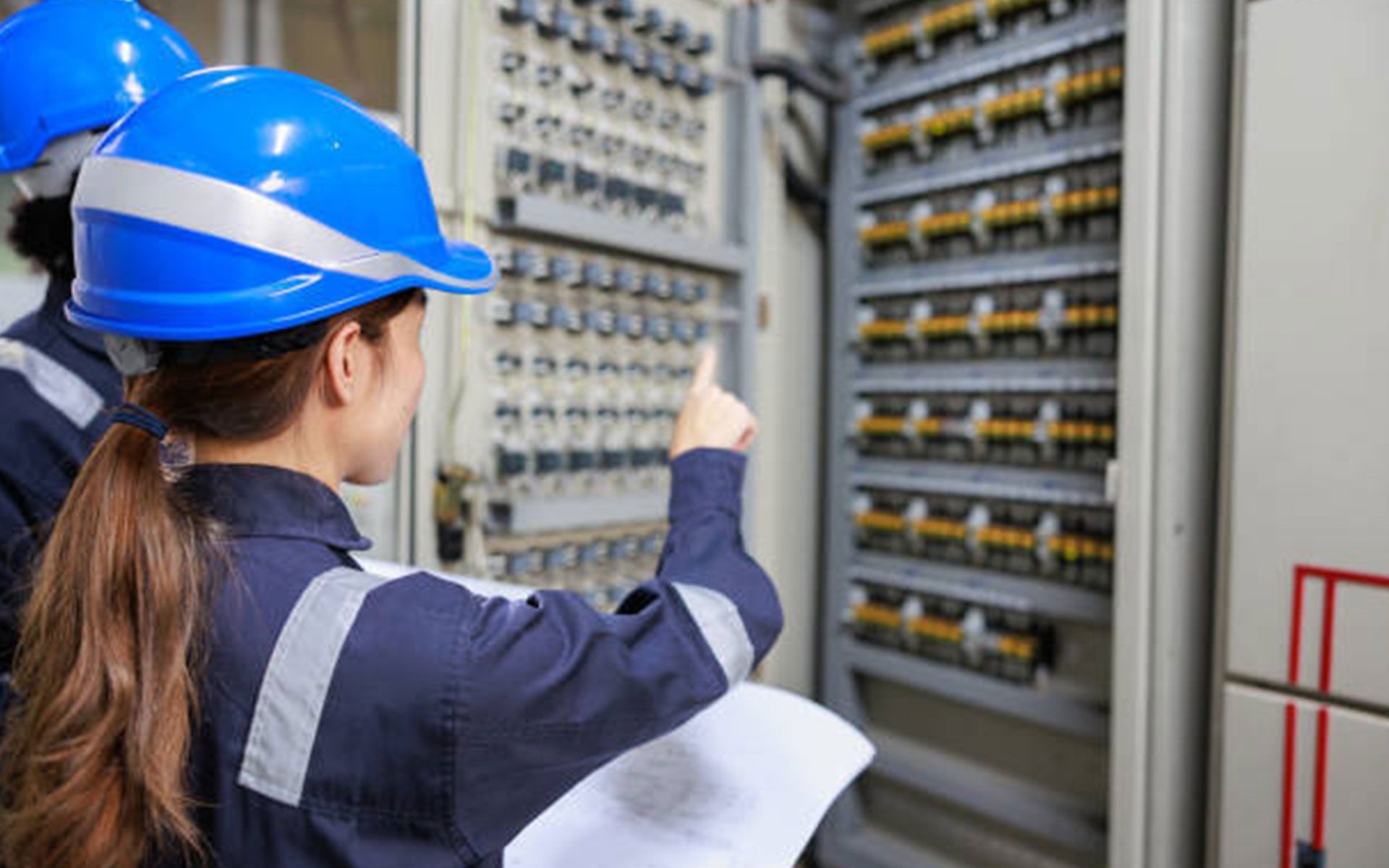 A woman in a hard hat is working on an electrical panel.
