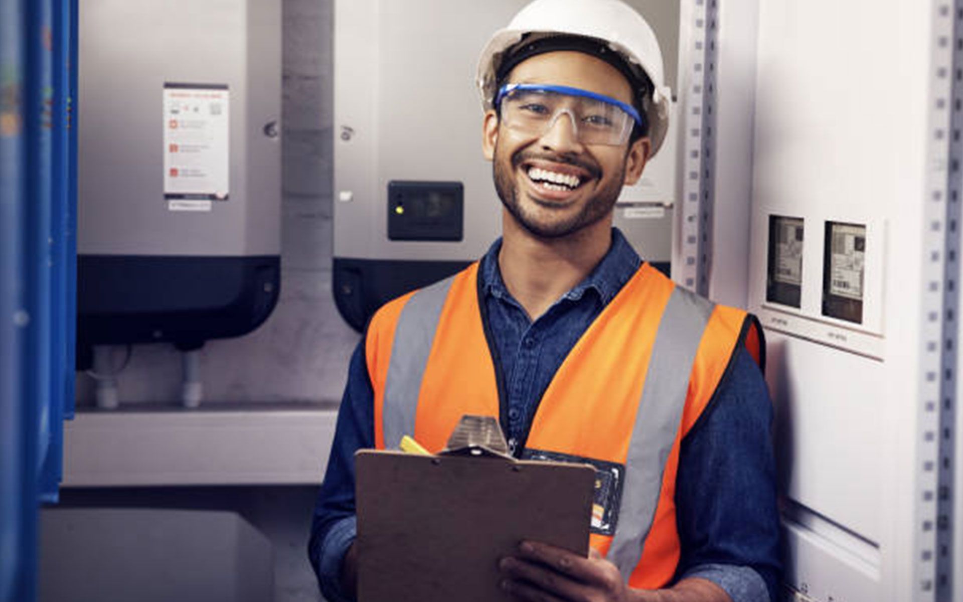 A man wearing a hard hat and safety vest is holding a clipboard and smiling.