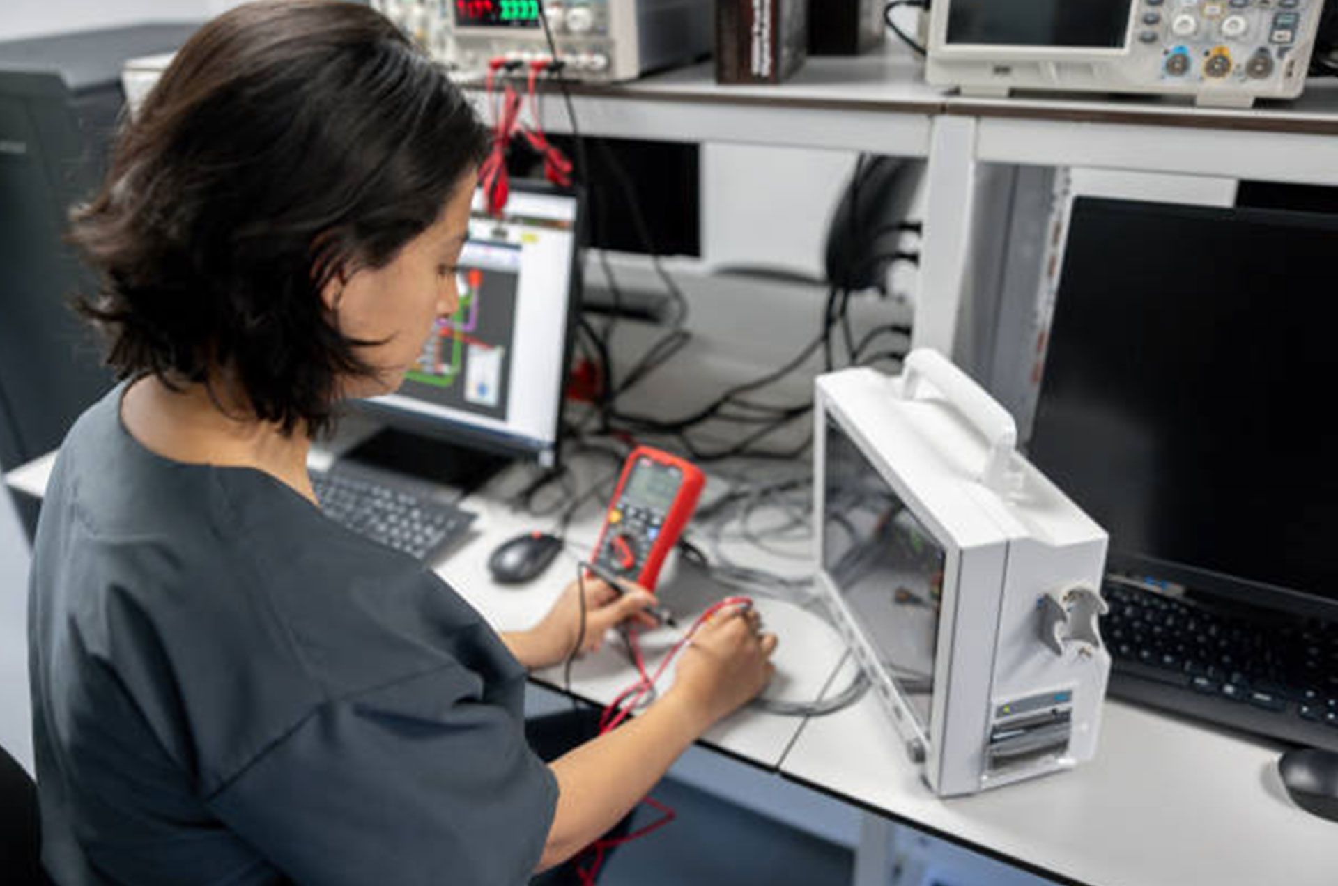 A woman is sitting at a desk using a multimeter.