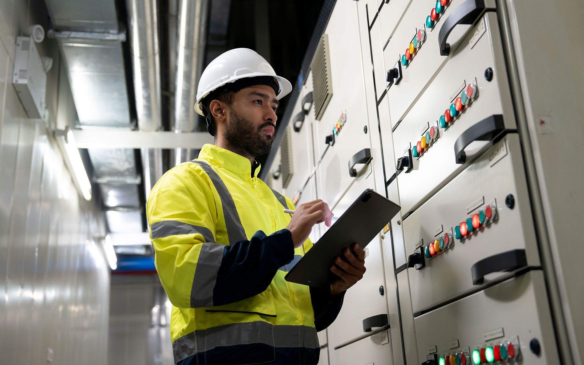 A man in a hard hat is holding a clipboard in front of a electrical panel.