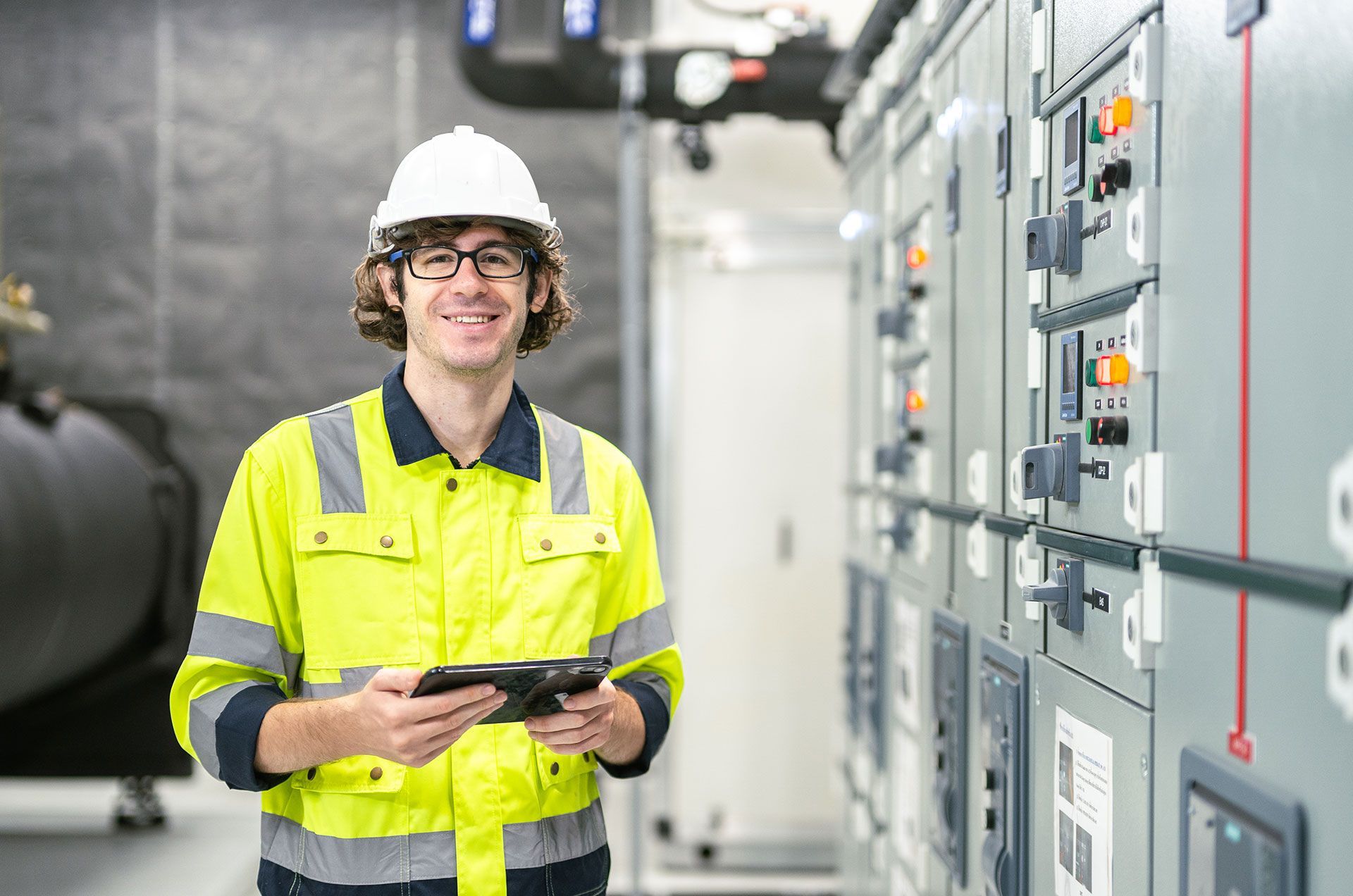A man in a hard hat is holding a tablet in a control room.