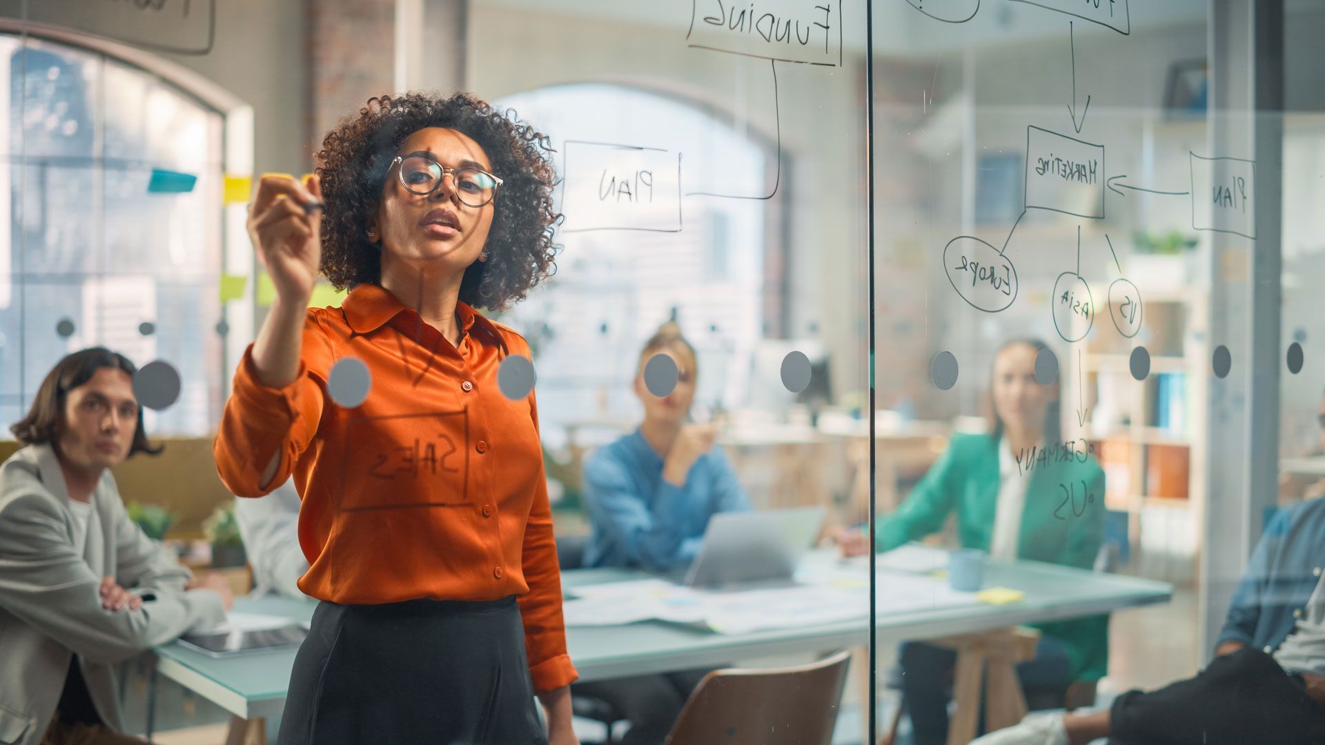 A woman is standing in front of a glass wall giving a presentation to a group of people.