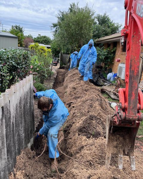 A Man in A Blue Jacket Is Digging in The Dirt Next to A Red Excavator — Northern Rivers Demolition & Asbestos in Ballina, NSW