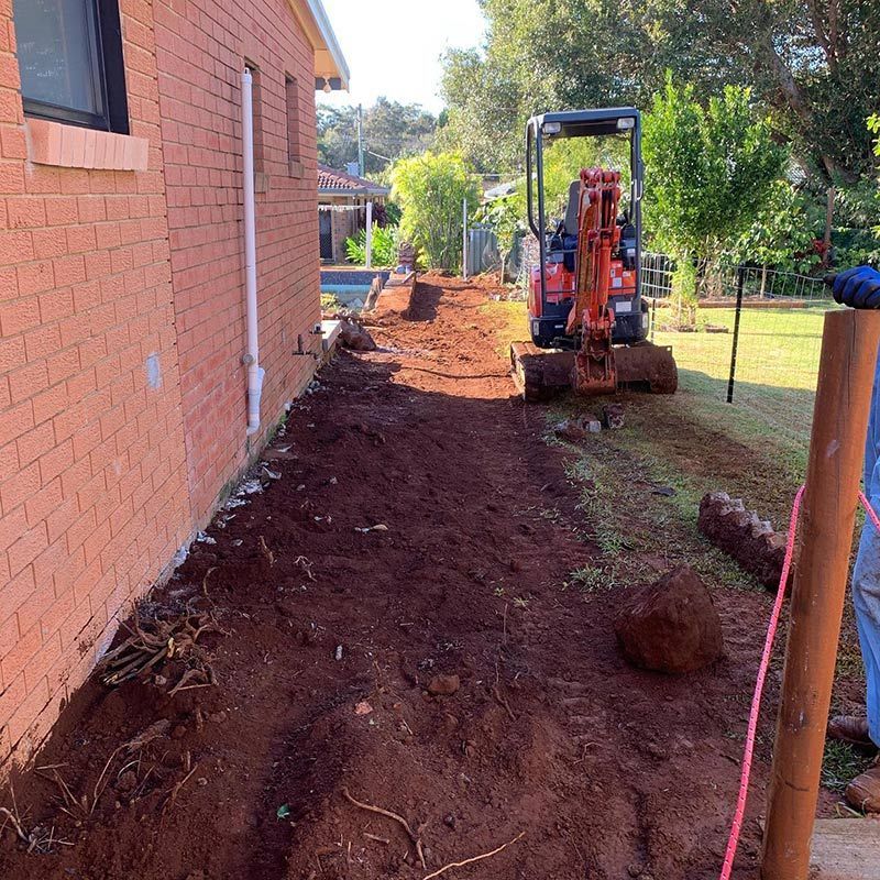 A Small Excavator Is Digging a Hole in The Dirt in Front of A Brick House — Northern Rivers Demolition & Asbestos in Skennars Head, NSW