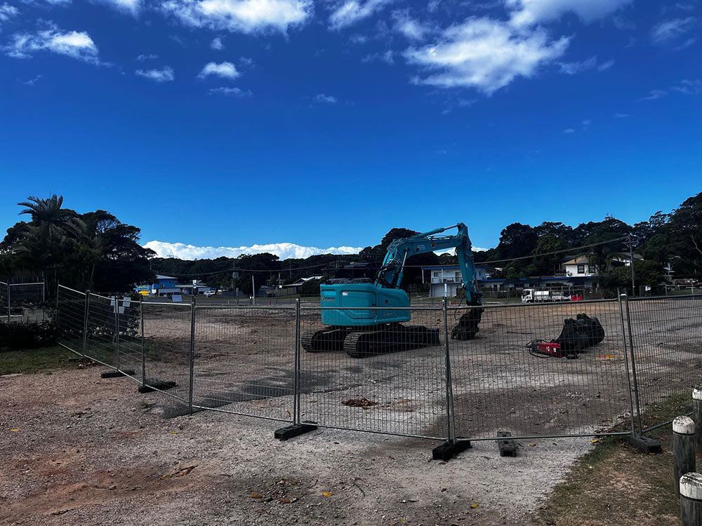 A Blue Excavator Is Parked in A Dirt Field Behind a Fence — Northern Rivers Demolition & Asbestos in Ballina, NSW