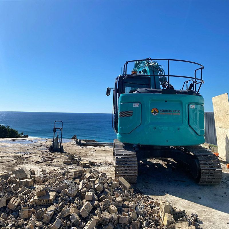 A Blue Excavator Is Parked on A Pile of Rocks Near the Ocean — Northern Rivers Demolition & Asbestos in Grafton, NSW