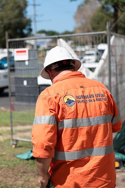 A Man Wearing a Hard Hat and An Orange Shirt Is Standing in Front of A Fence — Northern Rivers Demolition & Asbestos in Murwillumbah, NSW