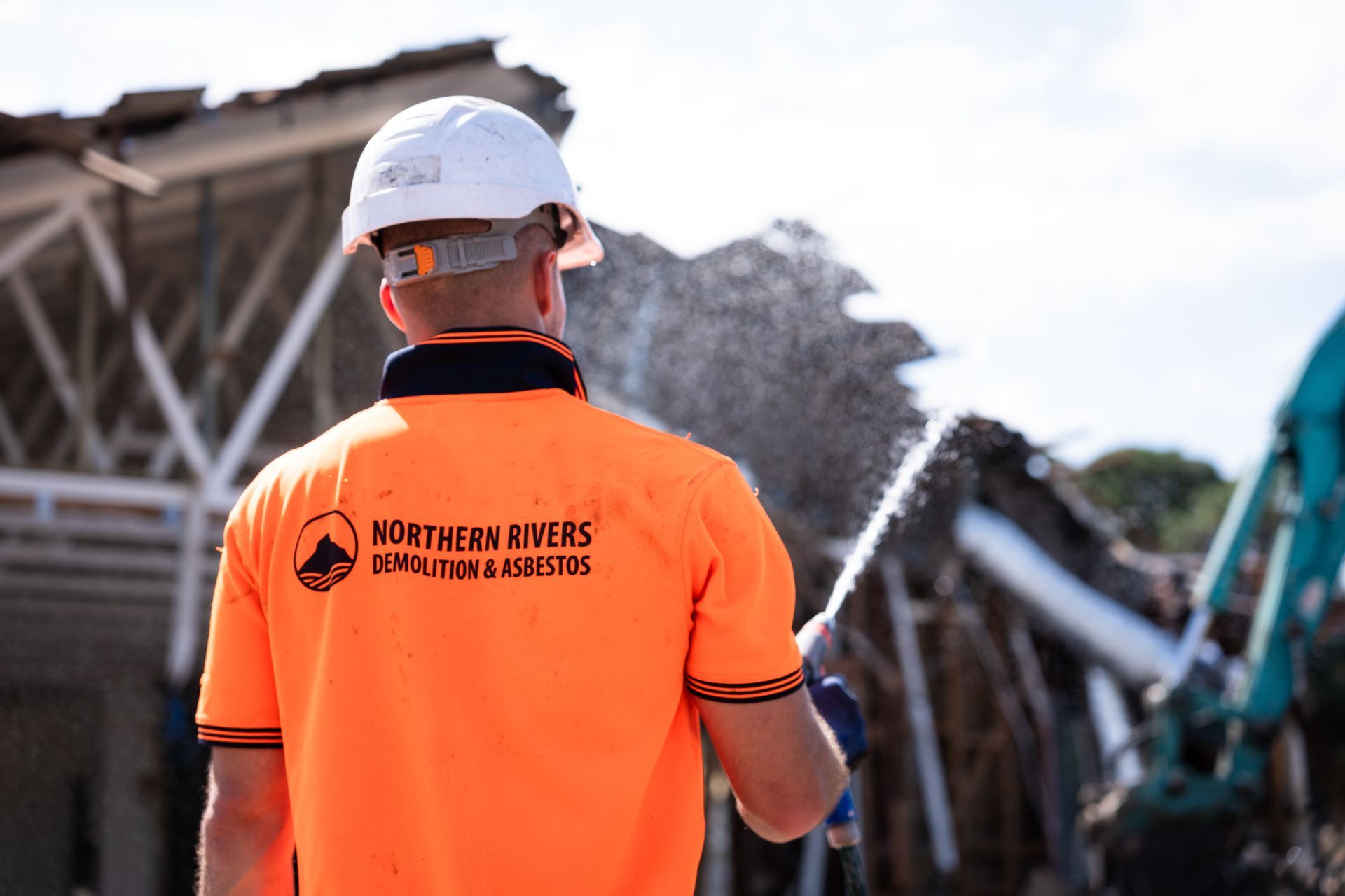 A man wearing a hard hat and an orange shirt is spraying water on a pile of rubble.