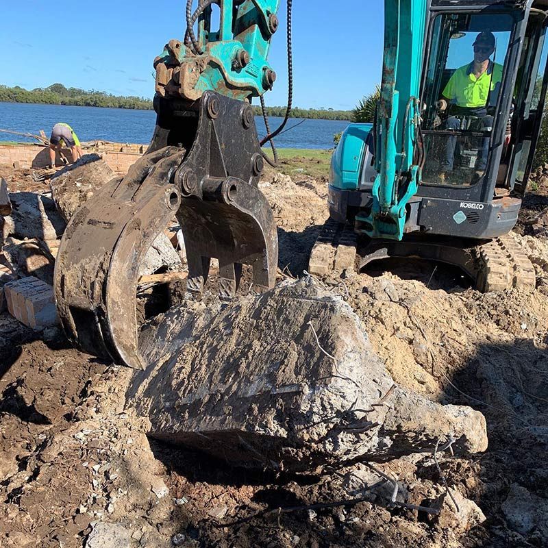 A Man Is Driving an Excavator in The Dirt Near a Body of Water — Northern Rivers Demolition & Asbestos in Yamba, NSW