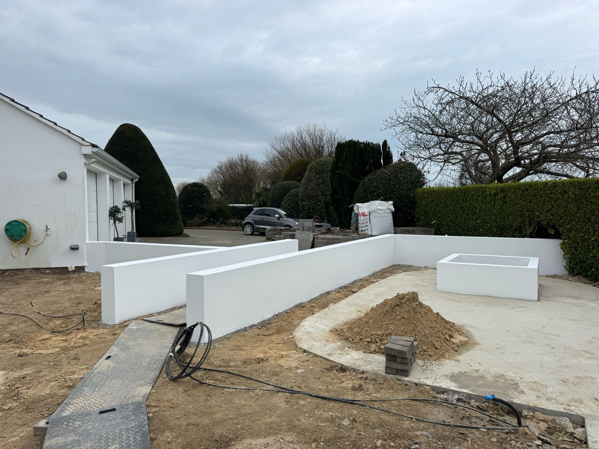 White walls and courtyard under construction, house in the background. Cloudy sky.
