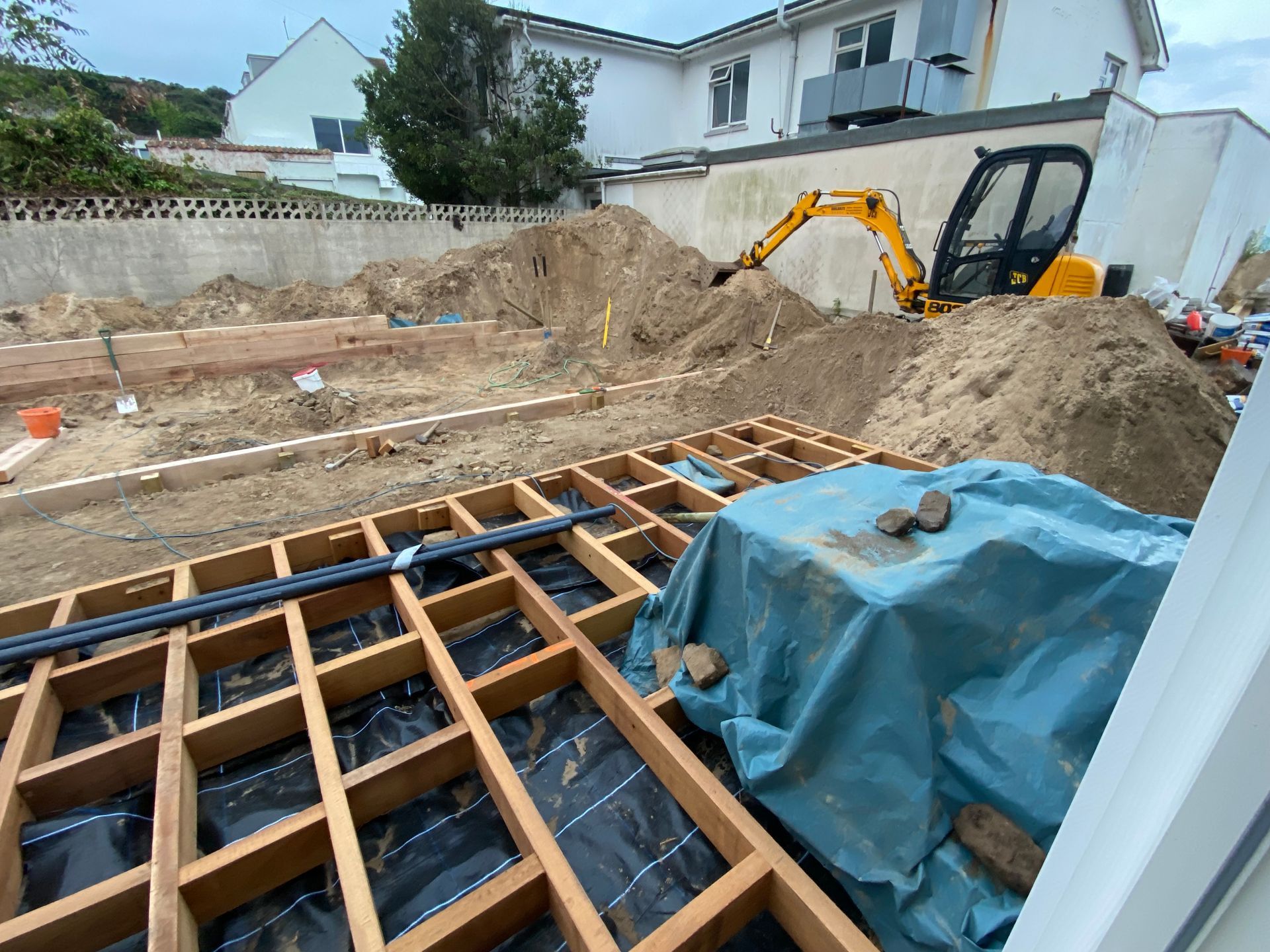 Construction site: wooden deck frame, excavator, piles of dirt, and building in the background.