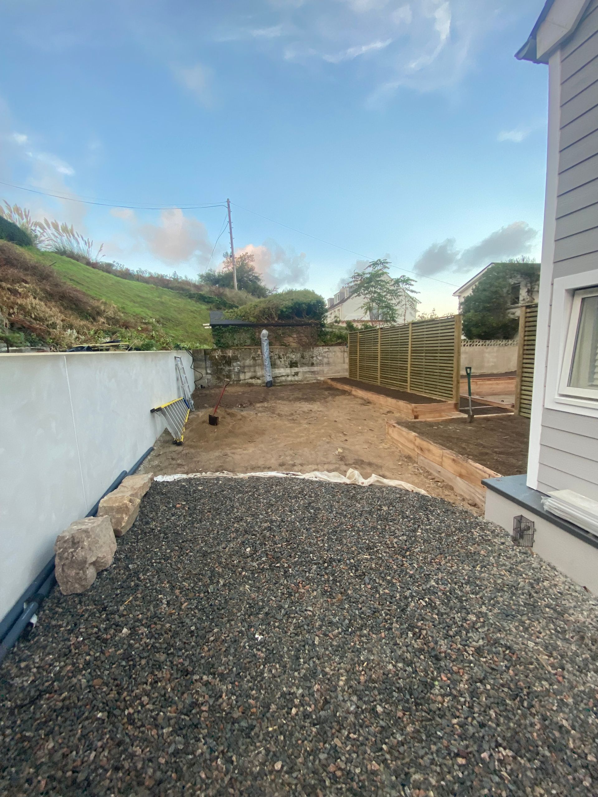 Gravel-covered yard with unfinished areas. White wall on the left, wooden structure at the back under a cloudy sky.