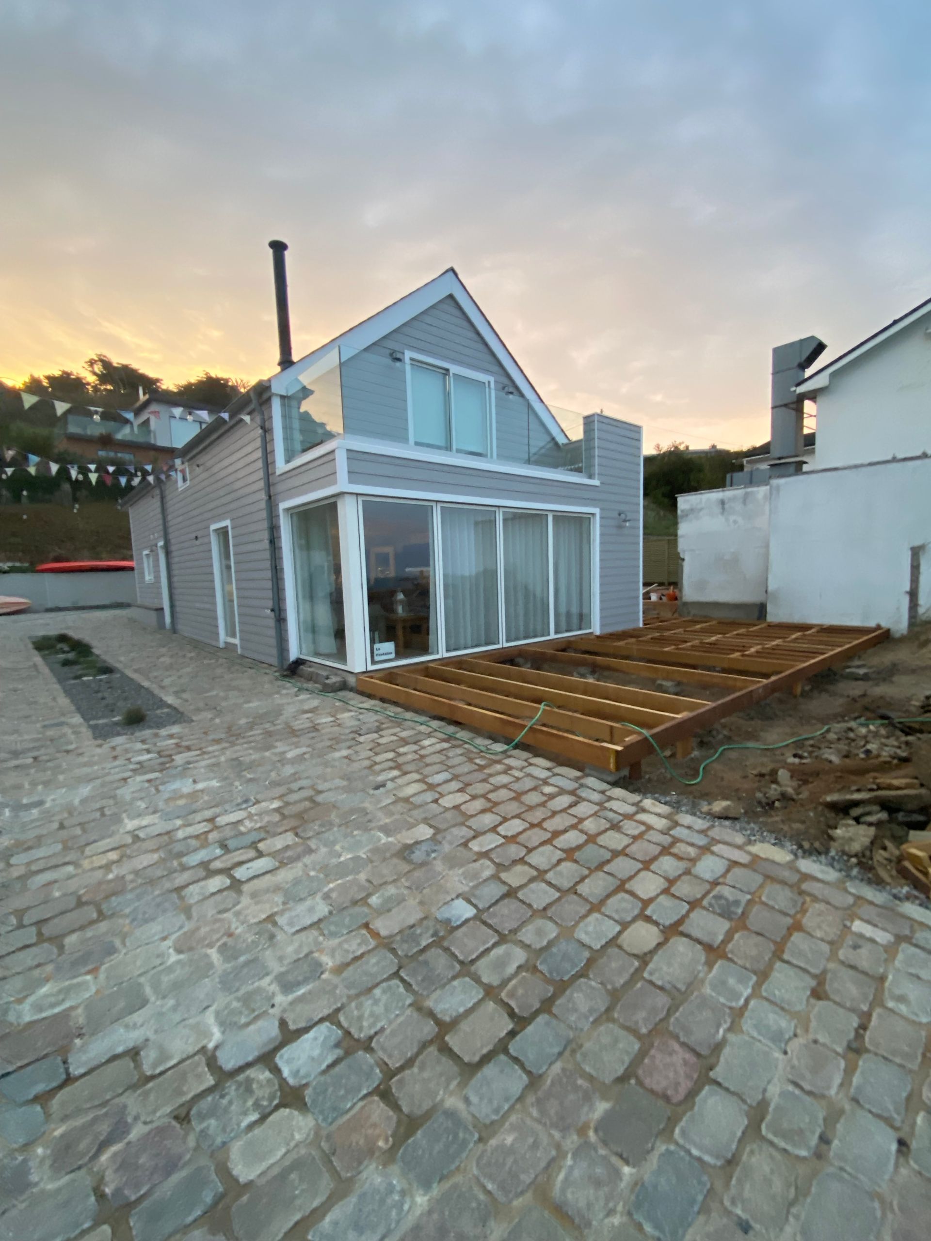 House with gray siding and glass windows, wooden deck under construction, cobblestone driveway.