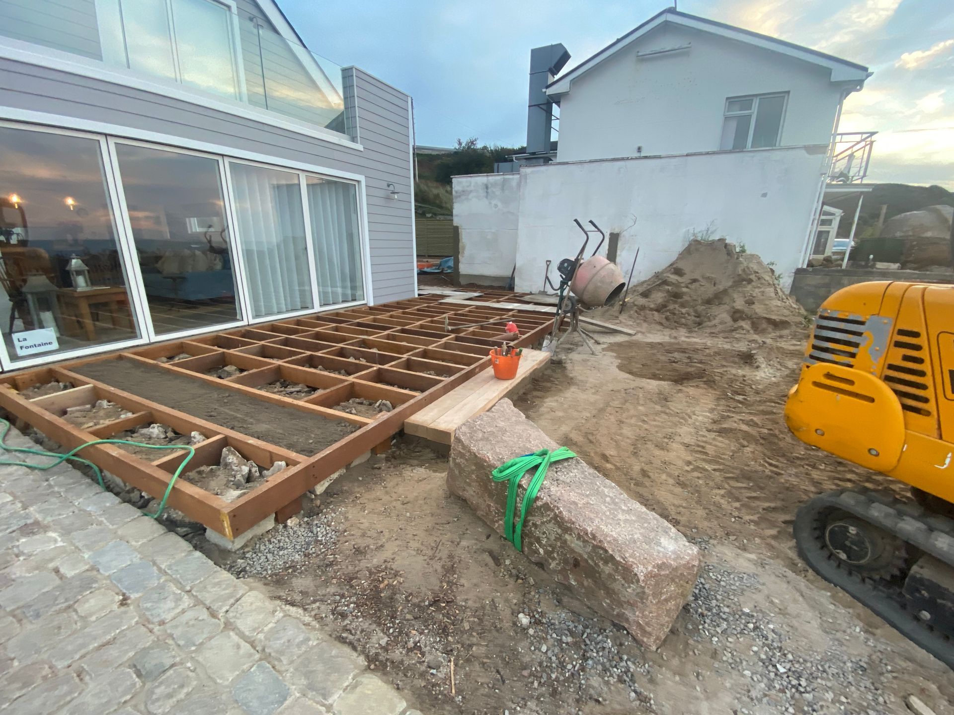 Deck construction: Wooden frame, soil, mini-excavator, cement mixer, house with sliding doors.