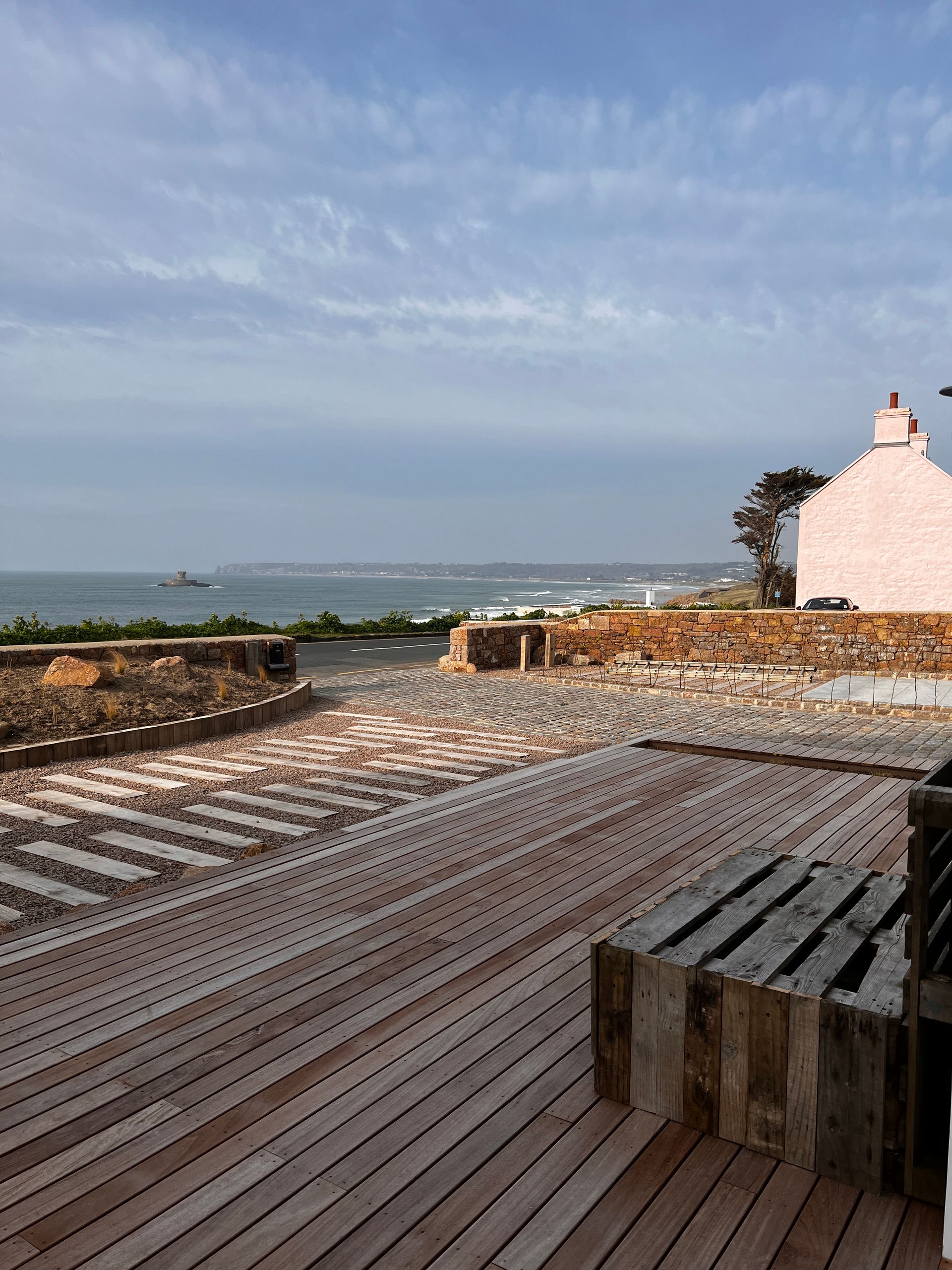 Wooden deck overlooking ocean, pathway, and pink building under cloudy sky.
