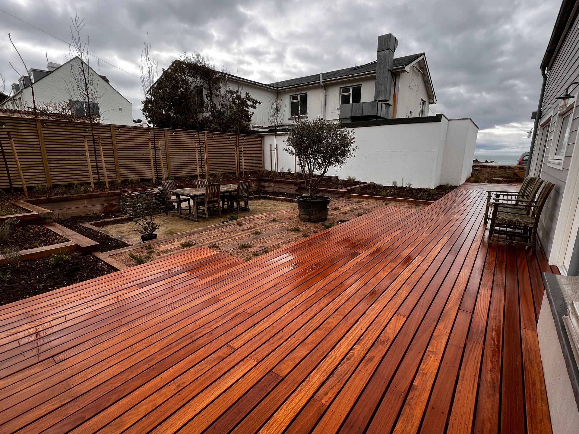 Wooden deck in a backyard with a seating area, brown and white buildings, cloudy sky.