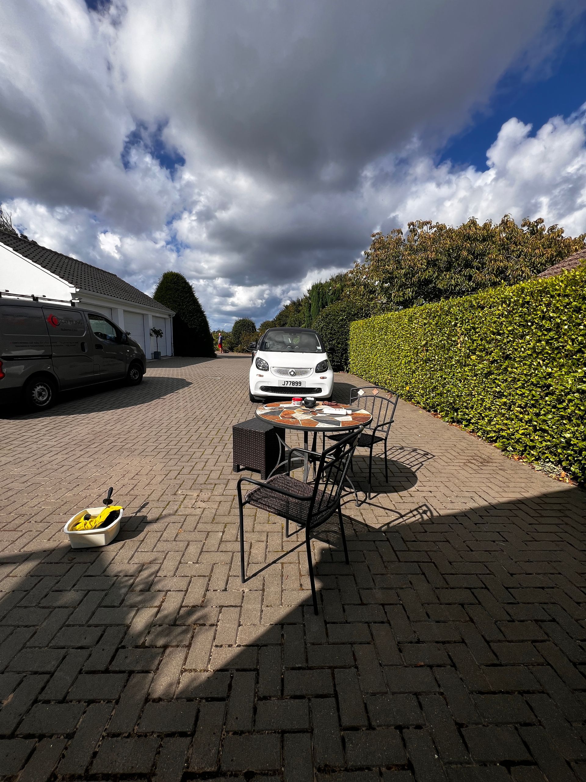 A white Smart car parked on a brick driveway, with a table set up in front of it. Overcast, sunny day.