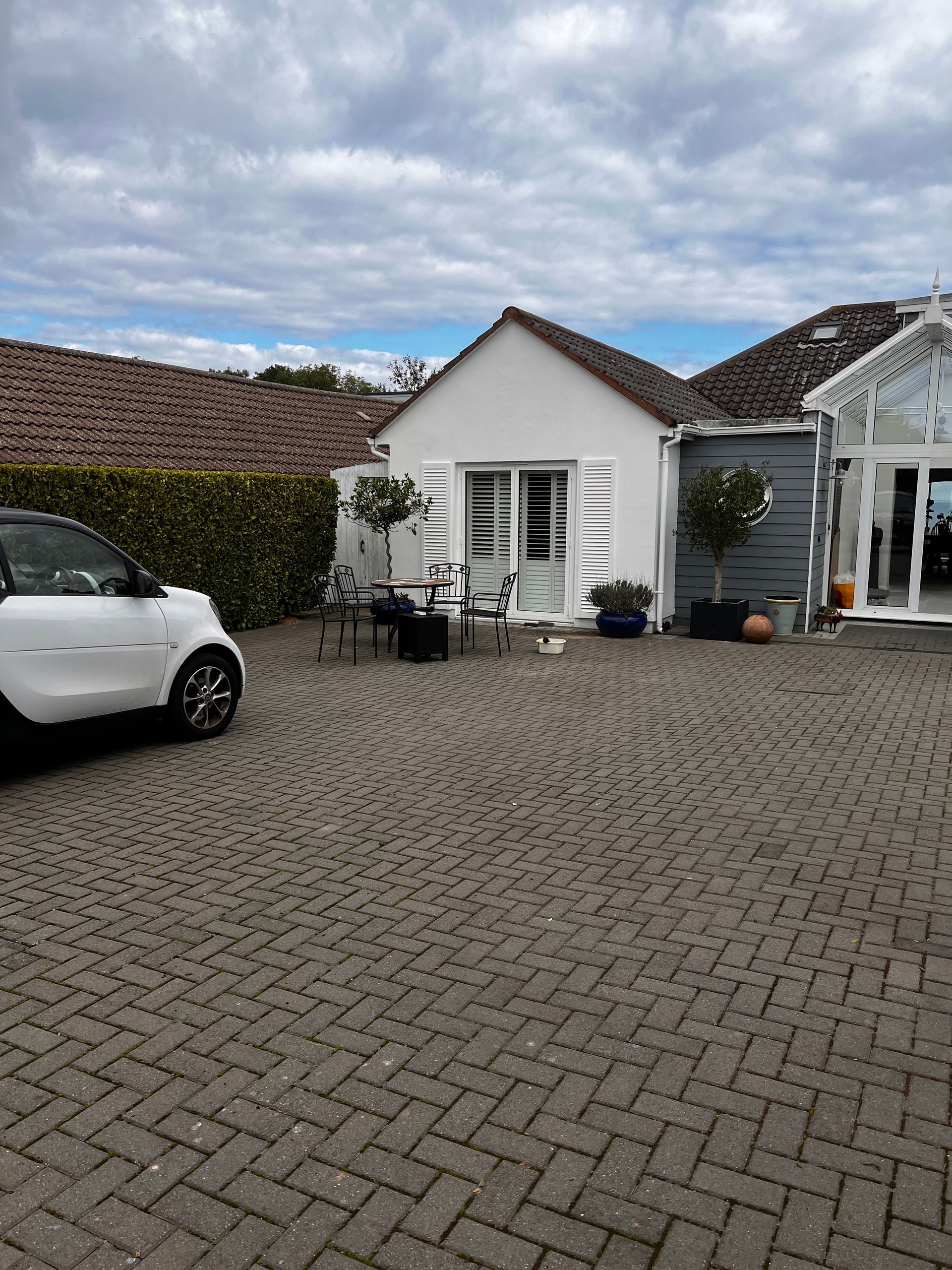 A white building with a small patio and a white car parked on brick pavers. Blue sky above.