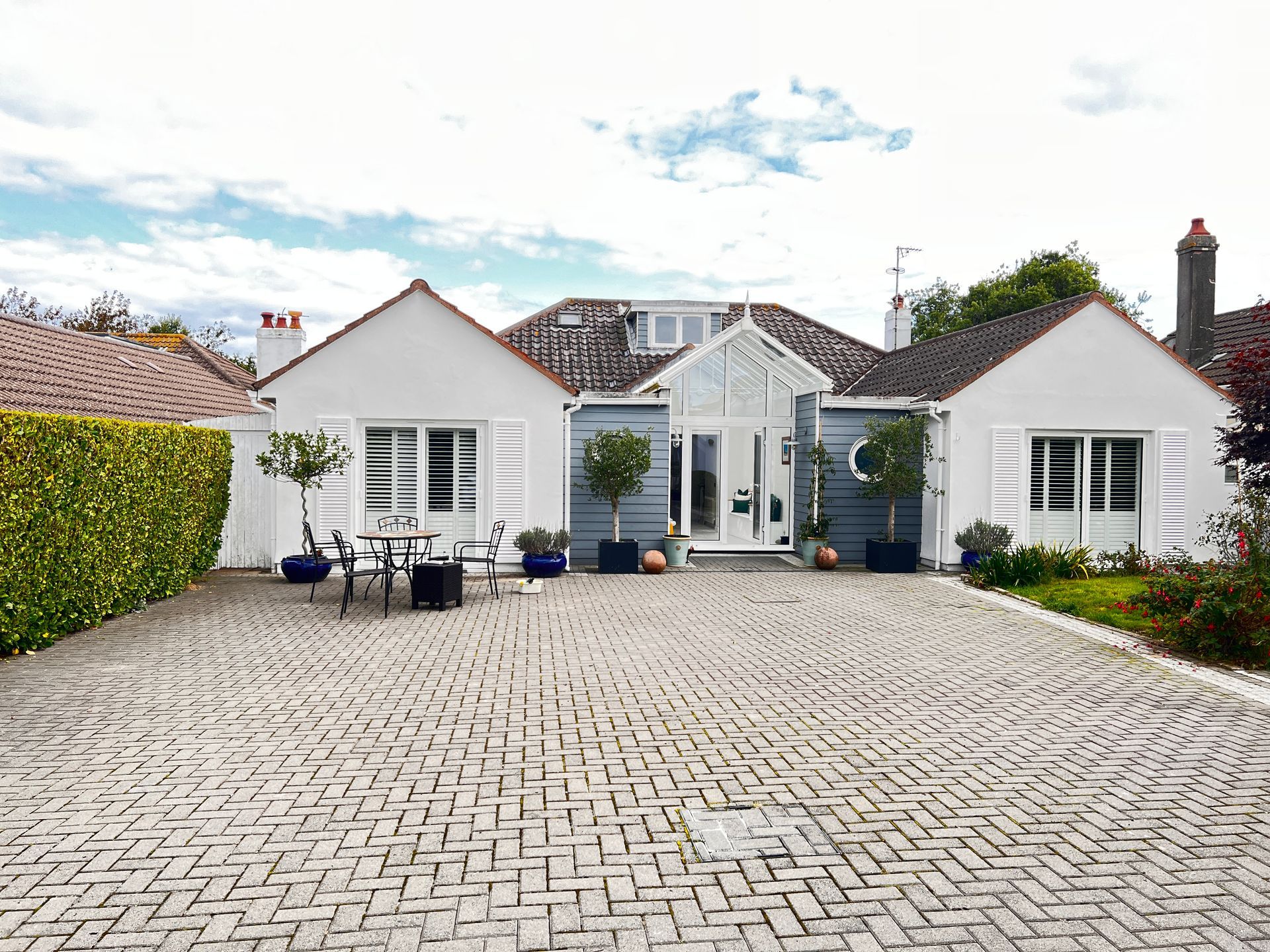 White house with brick driveway, shutters, small trees in pots.